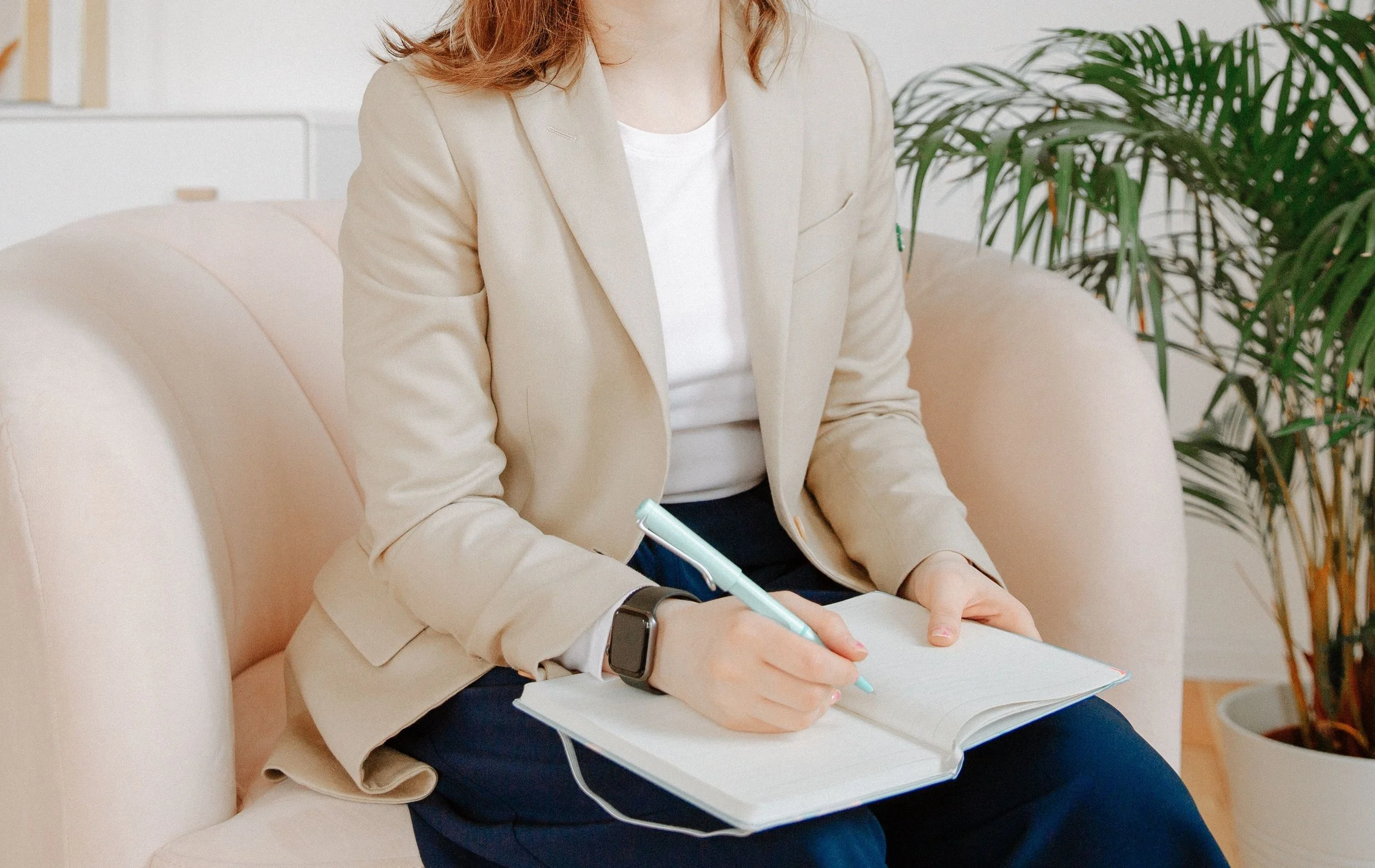 Close-up of a woman sitting on a light-colored sofa, wearing a beige blazer and white top, holding a notebook and a mint green pen, with a plant in the background.