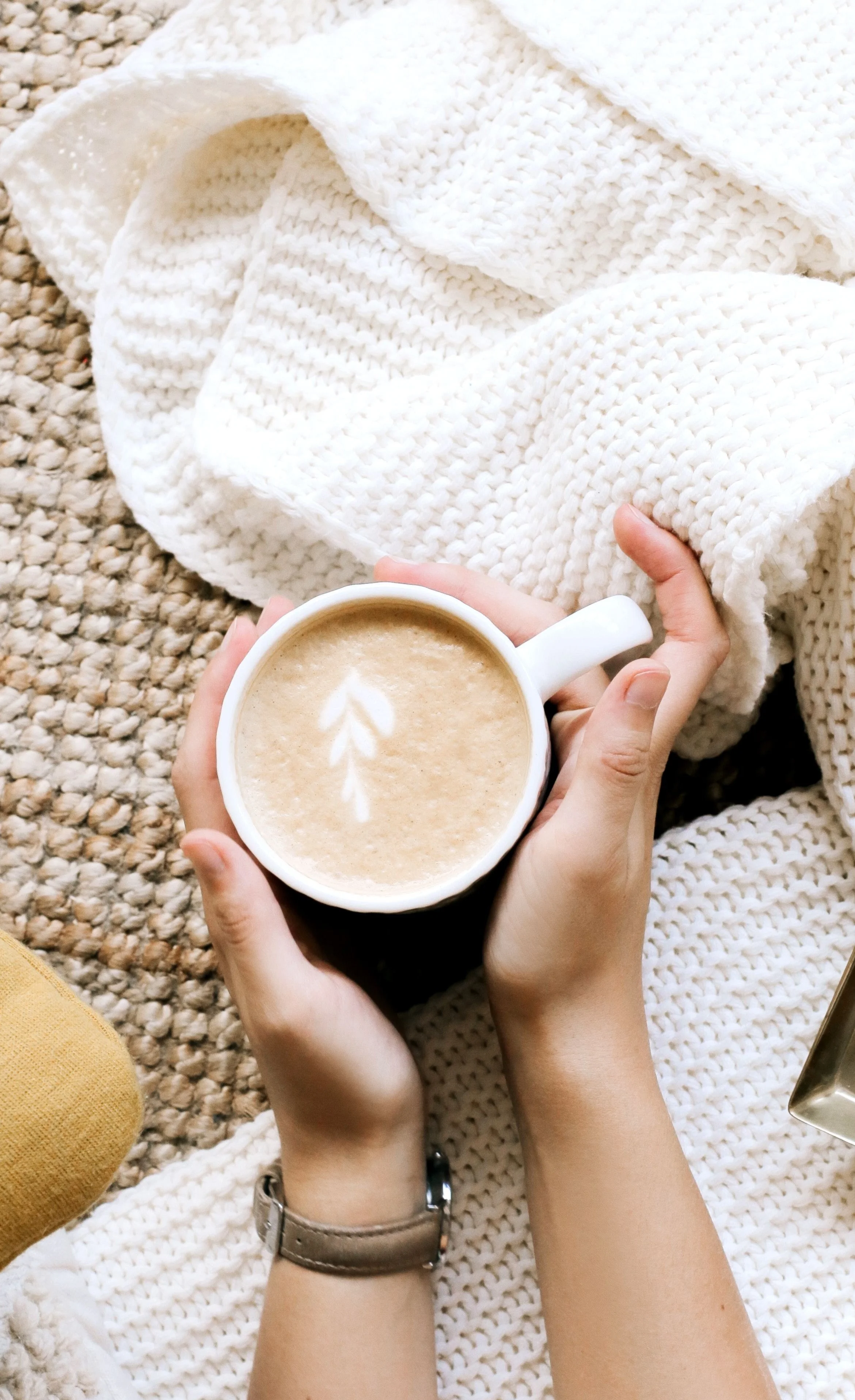 Person holding a white coffee mug with latte art, sitting on a chunky white knit blanket on a textured beige rug.