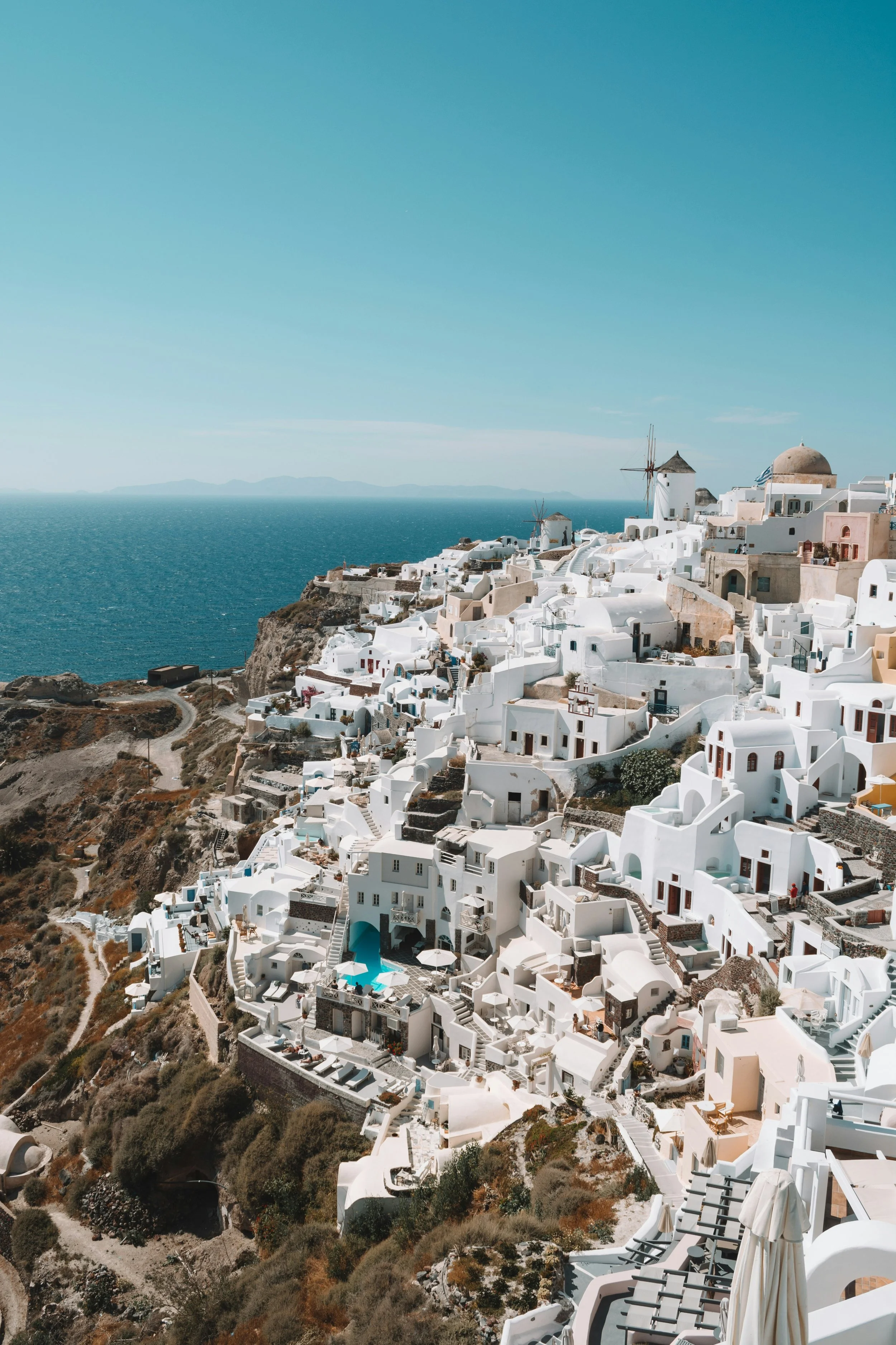 Aerial view of white buildings with domed roofs and a windmill on a cliffside in Santorini, Greece, overlooking the ocean and distant mountains under a clear blue sky.