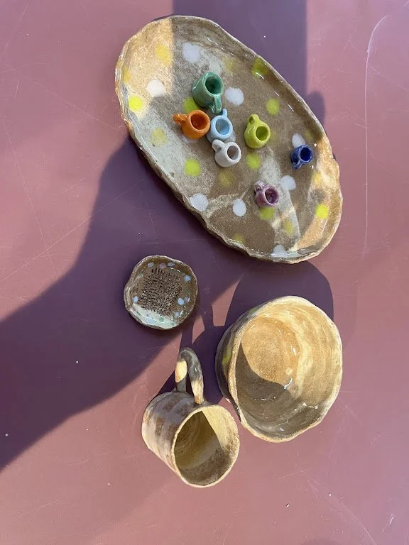 Een versierd bord en kopjes op een roze tafel, met een hand die in beeld is. De bordjes hebben verschillende kleuren en patronen.
