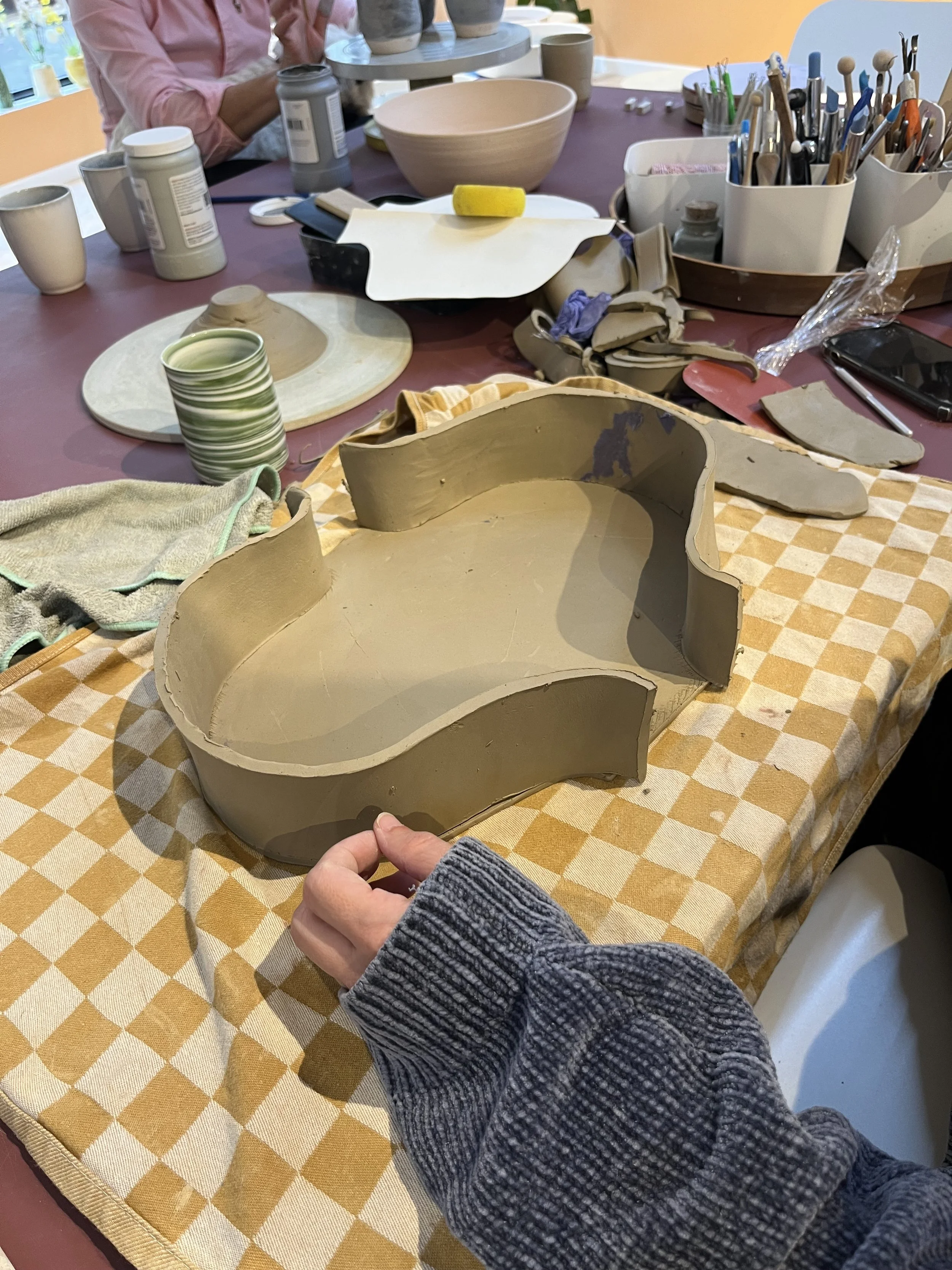 Persoon die een keramisch object vastpakt, omringd door keramische werkstukken en gereedschap op een tafel in een atelier.