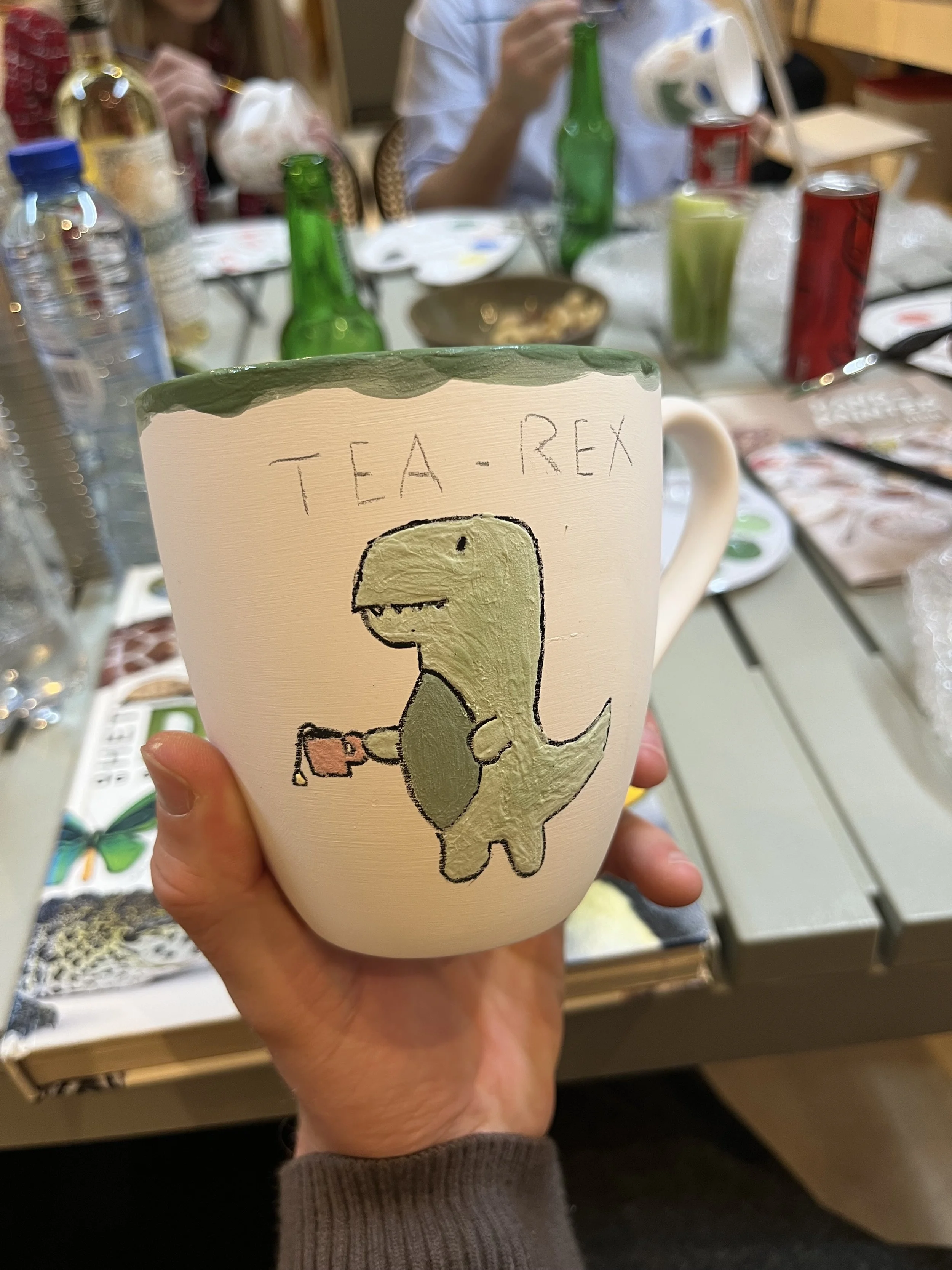 Een persoon houdt een aardig gedecoreerde witte beker vast met een tekening van een dinosaurus die een thee kopje vasthoudt en de woorden 'TEA-REX' erboven geschreven.