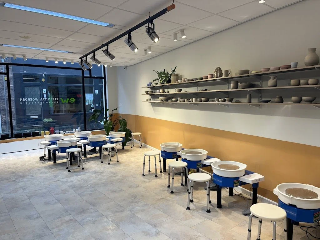 Binnenkant van een keramiekstudio met werkbanken, keramiekdraaibanken, planken met potten en vazen, grote ramen, en enkele planten.