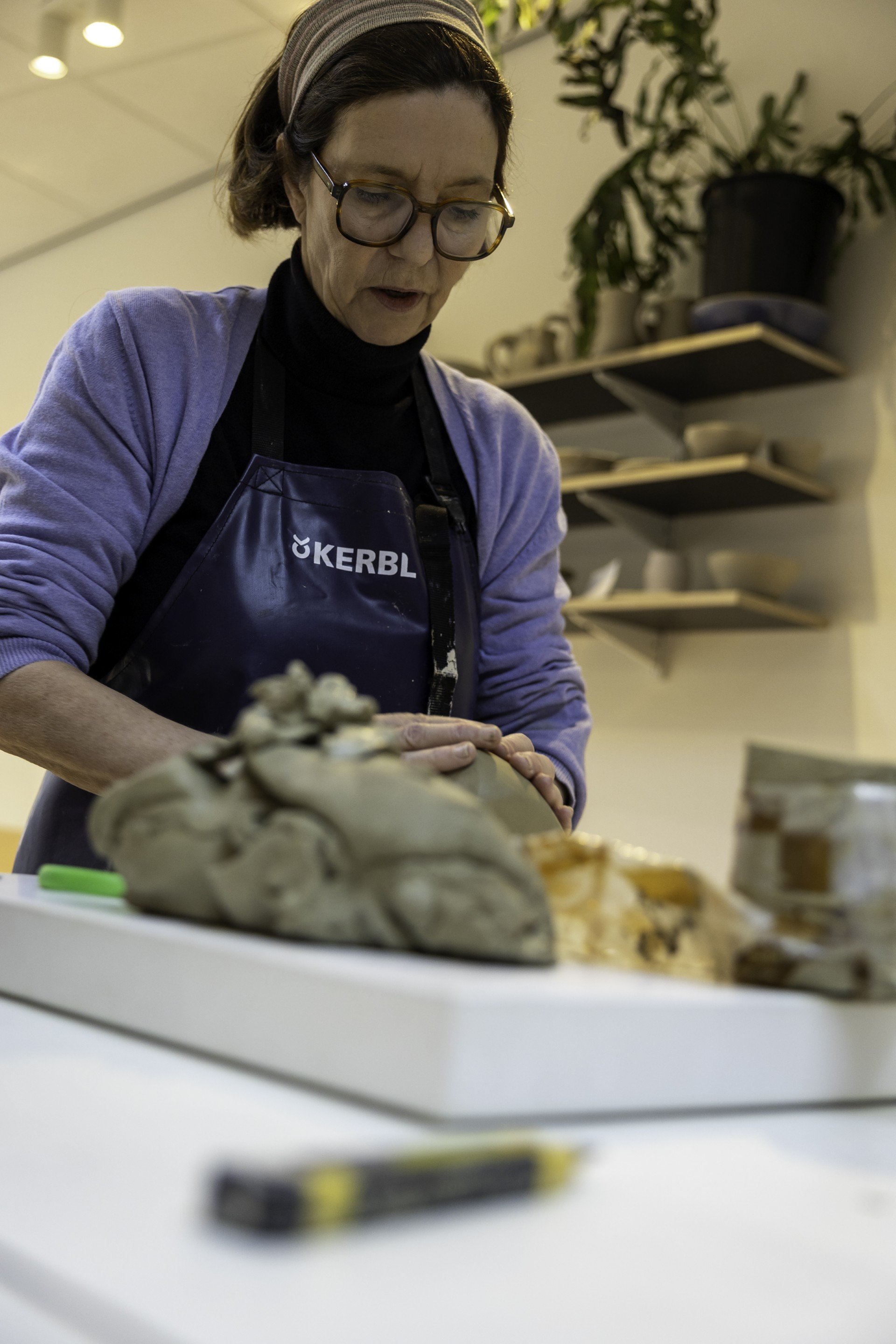 Een vrouw met bruin haar, grote bril, een hoofdband en een zwart schouderkraag met het logo 'KERBL', werkt aan een kunstwerk op een tafel. Op de achtergrond bevinden zich planten en planken met potten en keramiek. Ze lijkt geconcentreerd te zijn op haar creatie.