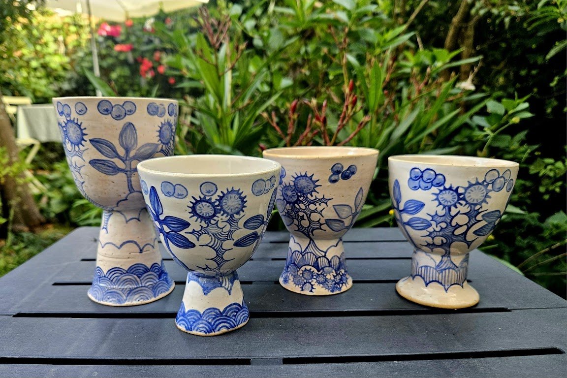 Vijf witte en blauwe keramische theekopjes met bloem- en bladmotieven, geplaatst op een zwarte houten tafel in een tuin.