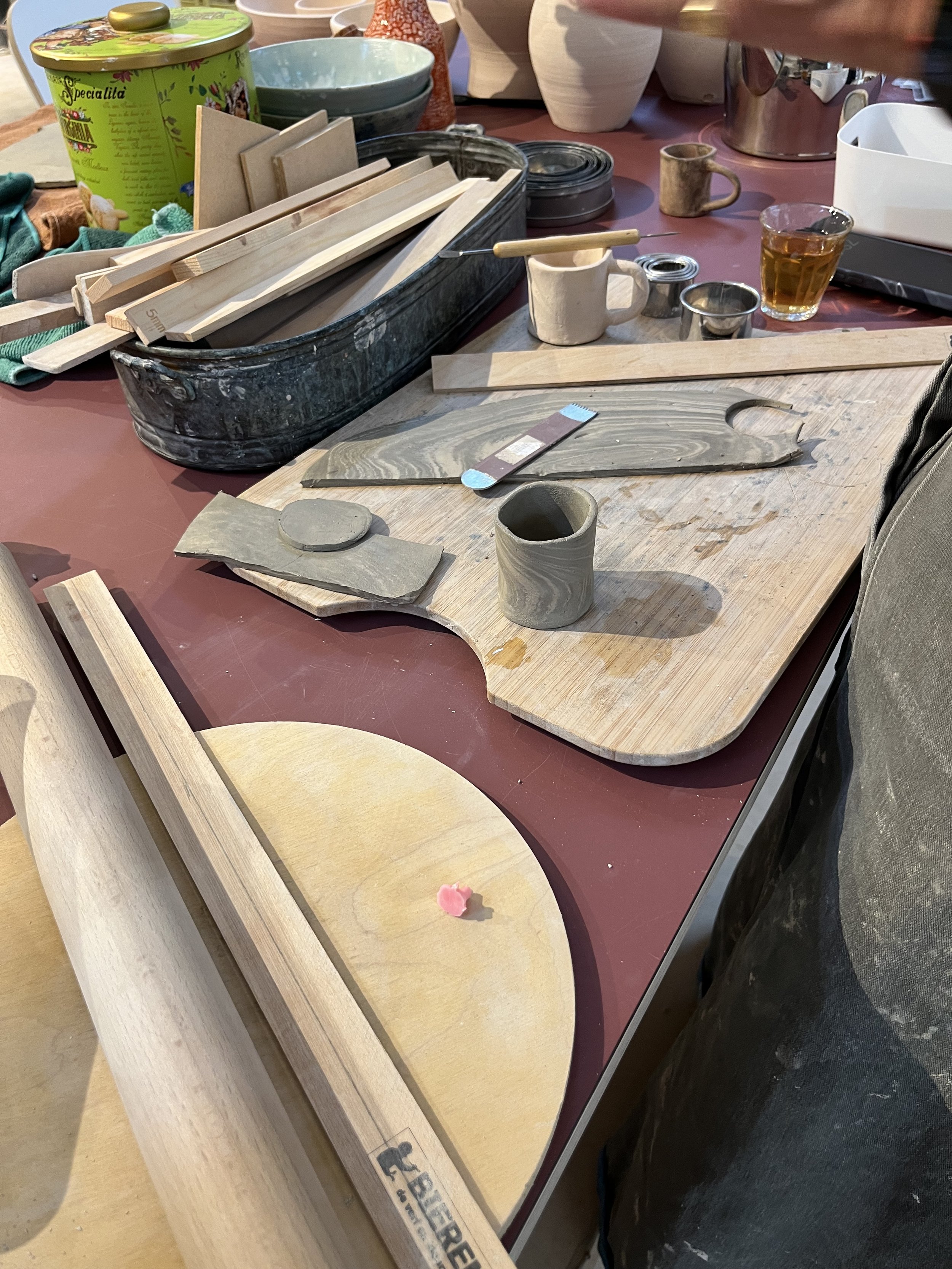 Werkplek voor keramiek of houtbewerking met houtplaten, gereedschap, keramiekwerkstukken, en diverse keramiek en gereedschap op een tafel.