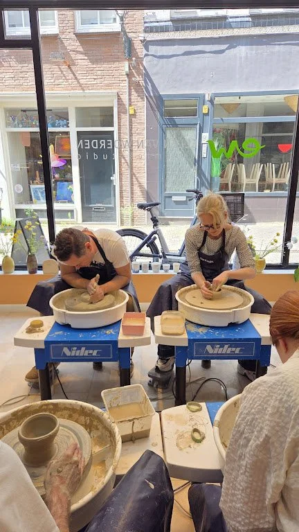 Twee mensen die pottenbakken maken op een keramiekdraaischijf in een gezellig atelier met grote ramen en uitzicht op straat.
