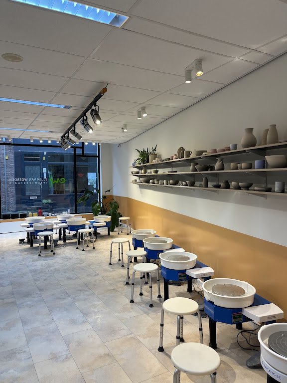Pottenbakkerij met keramiektafels en een muur met open planken gevuld met keramische vazen en kommen, grote ramen aan de voorkant, moderne inrichting.