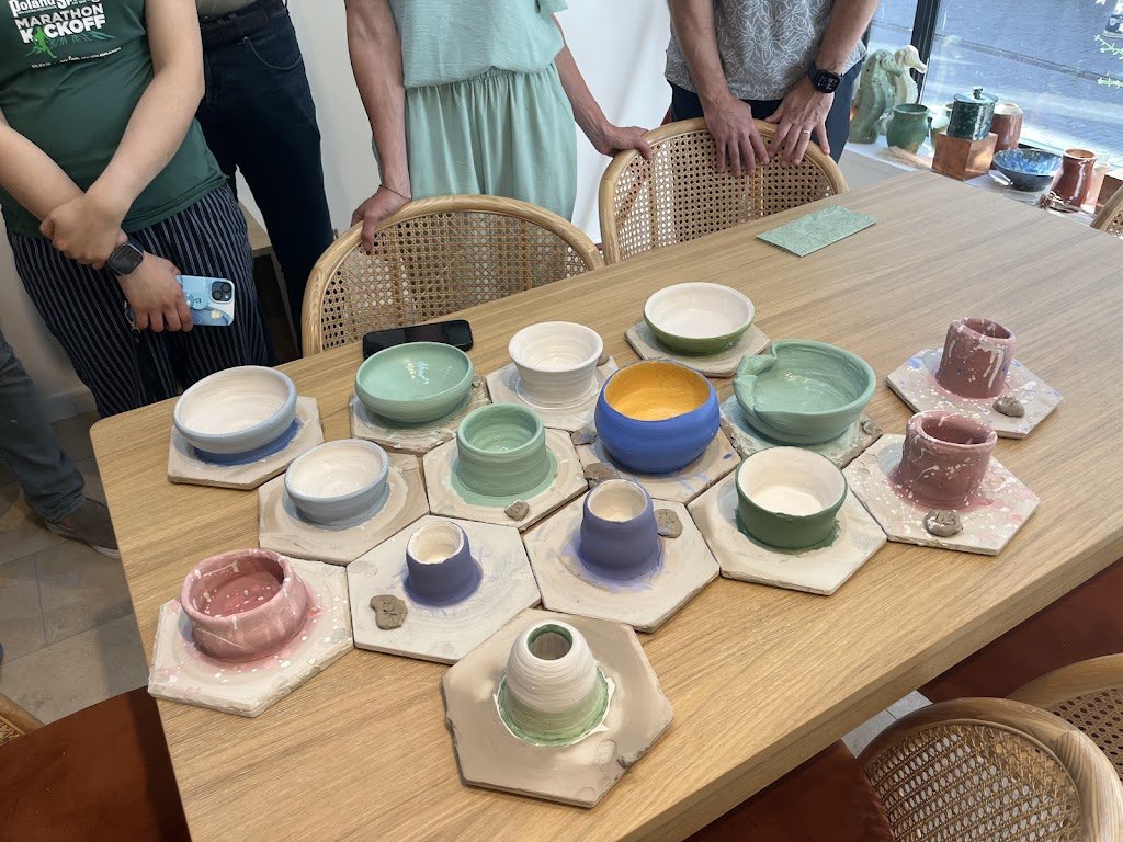 Een tafel met verschillende handgemaakte keramieken potten en kommen in diverse kleuren en vormen, omgeven door mensen die eromheen staan.