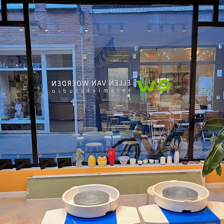 Binnen en buiten uitzicht via grote winkelraam met keramische potten, stoelen, en tafels.