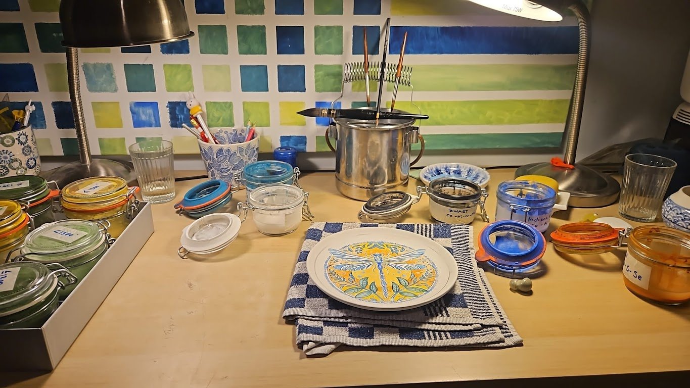 Een keukenwerkbank met diverse potjes en borden, een kopje met potloden en verfkwasten, een grote pan met prikkers, en een bord met een ontwerp met een libelle en bloemen.