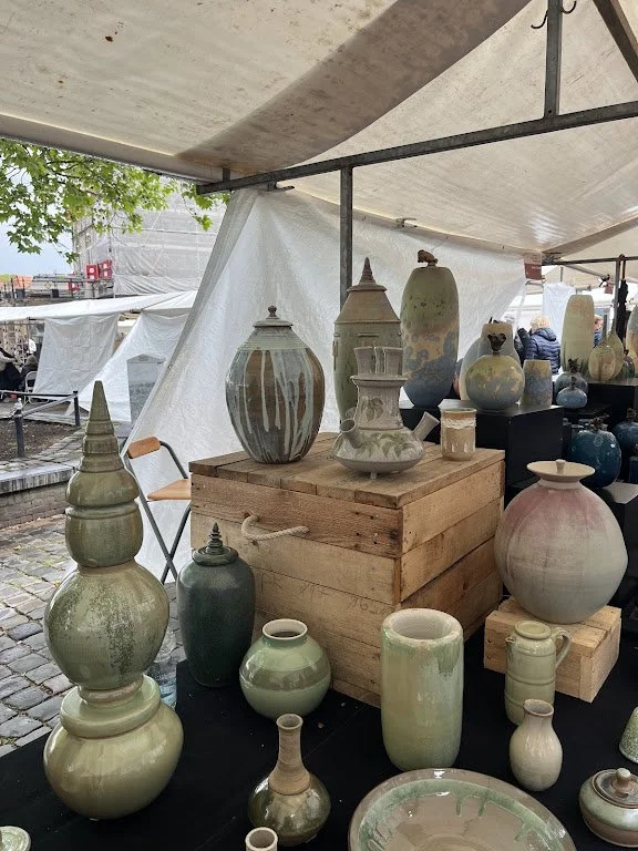 Potterie tentoongesteld onder een overkapping op een markt, met verschillende groottes en vormen, in aardse tinten.