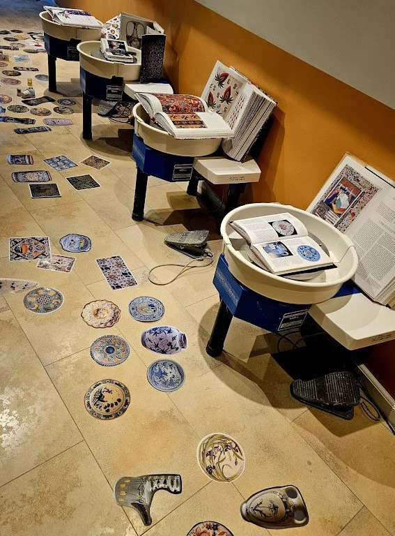 Tafels en boekenrekken met open boeken en decoratieve bordjes die op de vloer liggen, in een winkel of bibliotheek met oranje muur.
