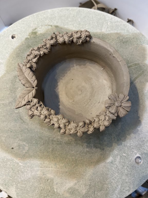 Een onvoltooide keramische kom met decoratieve bloem- en bladmotieven die rond de rand zijn aangebracht.