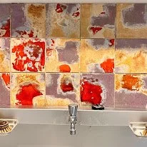 Saillant muur van gekleurde stenen met een rood, oranje en paarse abstracte verfpatroon boven een wastafel met twee schaaltjes.