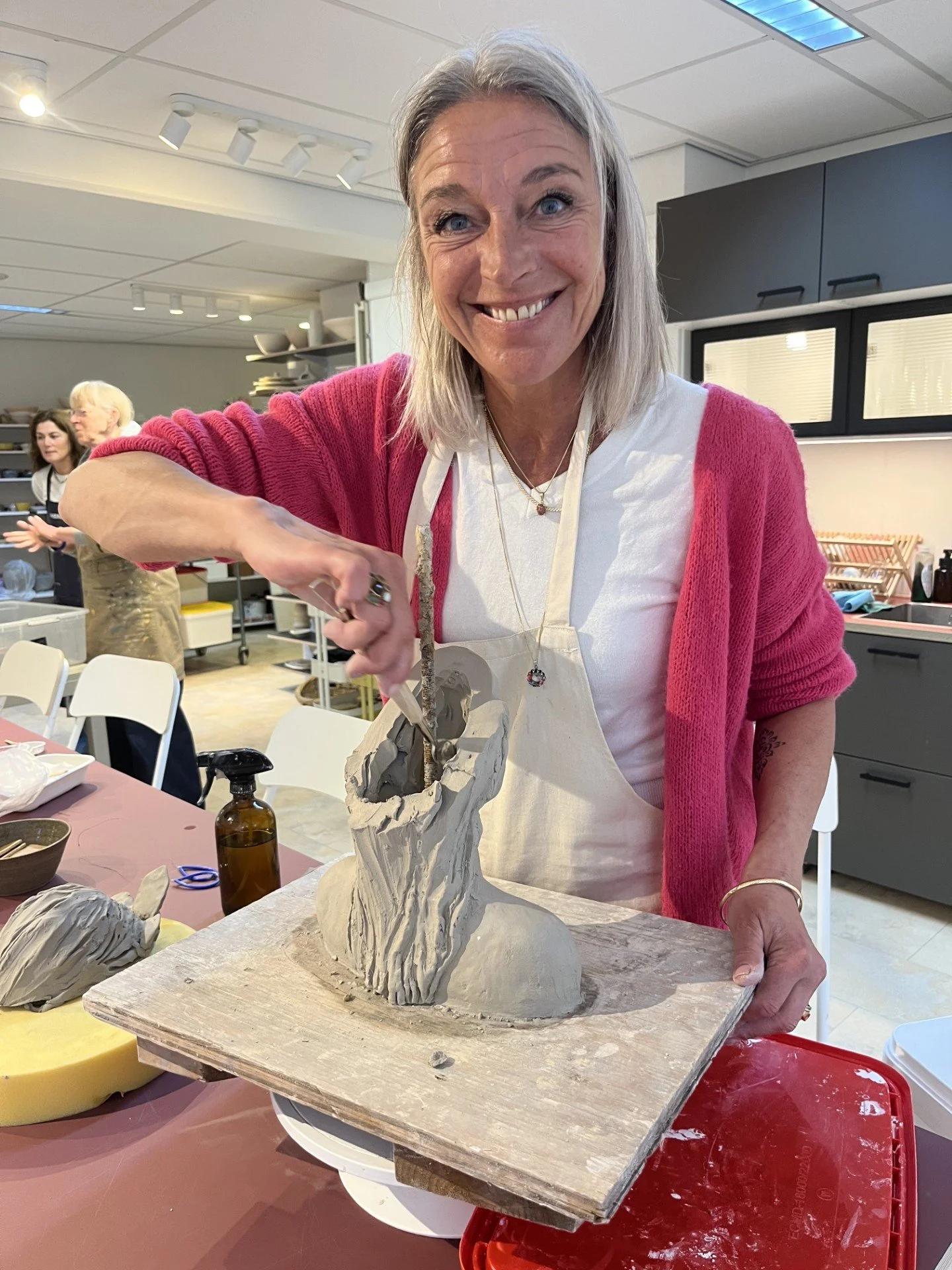 vrouw die een keramisch beeldhouwwerk maakt in een keuken of atelier, met andere mensen op de achtergrond
