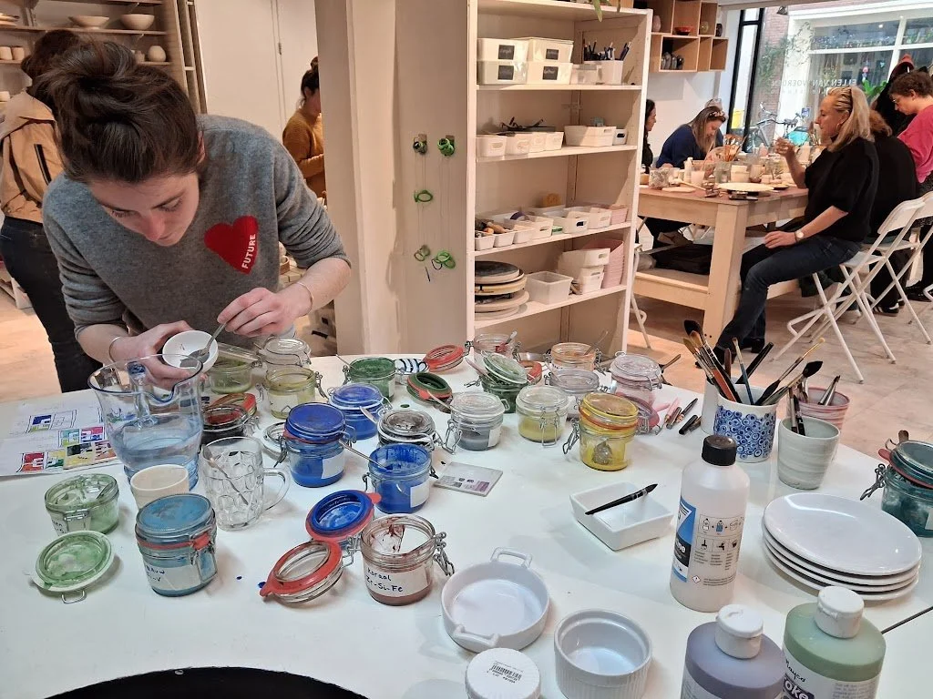 Een groep mensen in een keramiekwerkplaats, met een vrouw die verf aan het mengen is op een tafel vol potjes met verf en keramieke voorwerpen, anderen in de achtergrond bezig met keramiek en schilderwerk.