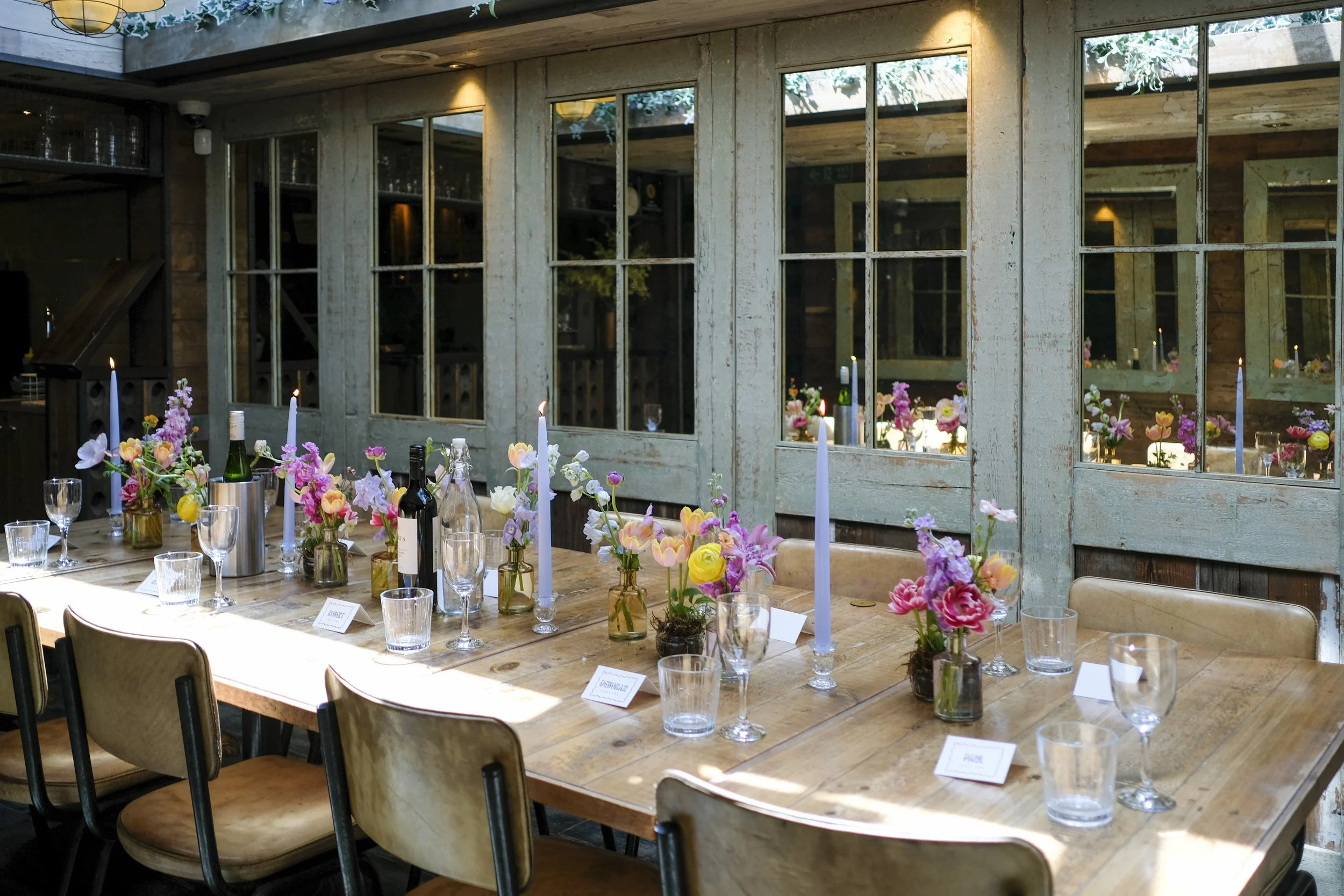 Interior of a rustic restaurant or event space with a wooden table decorated with colorful flower arrangements, wine bottles, glasses, and candles, set against a backdrop of large windows with a view of a wooden ceiling and natural light.