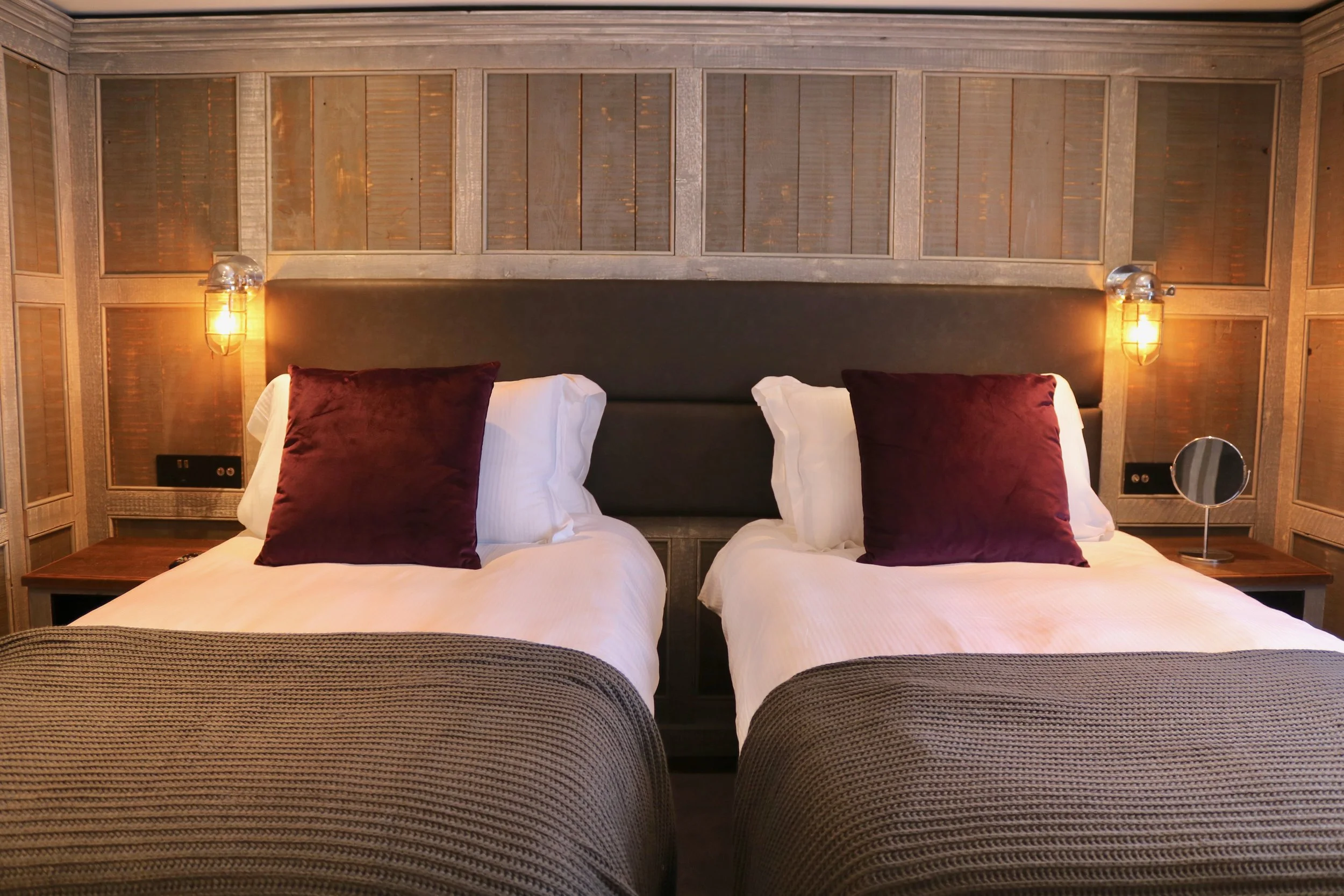 Two beds in a hotel room with dark headboards, burgundy pillows, white linens, and textured brown blankets. Behind the beds, a decorative wooden wall with two wall-mounted lamps on either side.