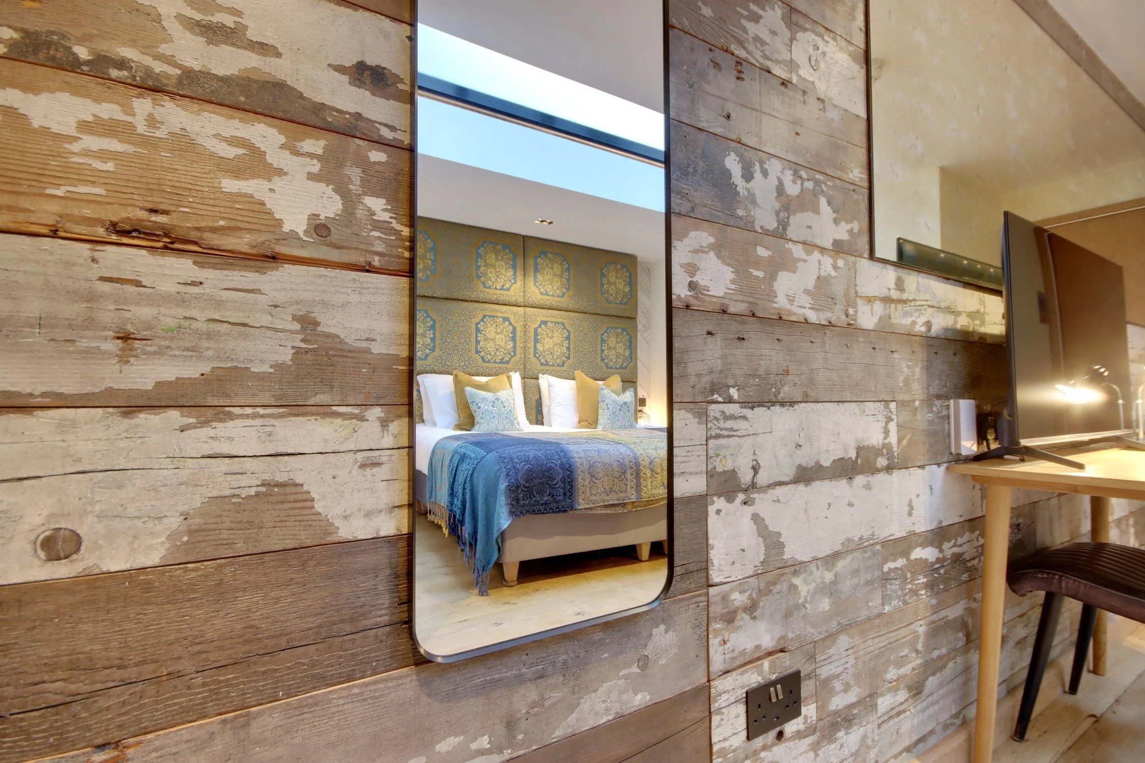Inside a room with a rustic wooden wall, a mirror reflects part of a bed with blue, yellow, and white bedding, a headboard with a decorative pattern, and ceiling with a skylight.