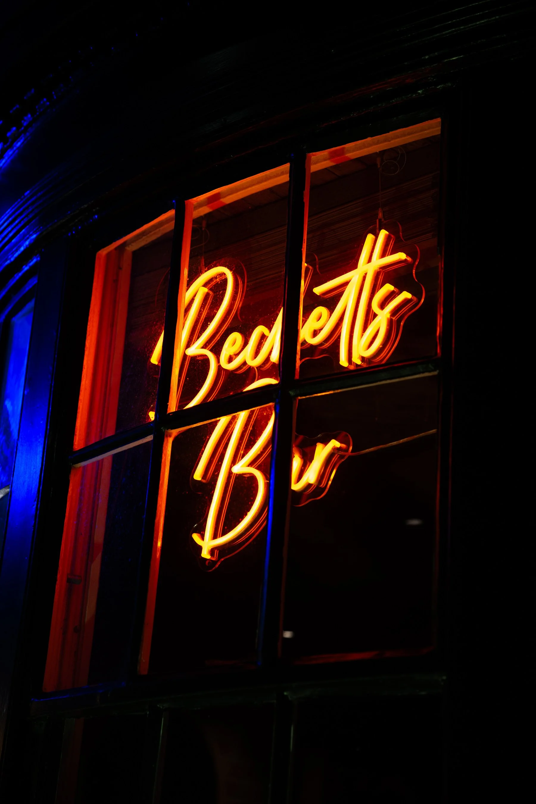 Neon sign that reads 'Betts Bar' in orange and yellow colors, displayed inside a window with black framing, against a dark background.