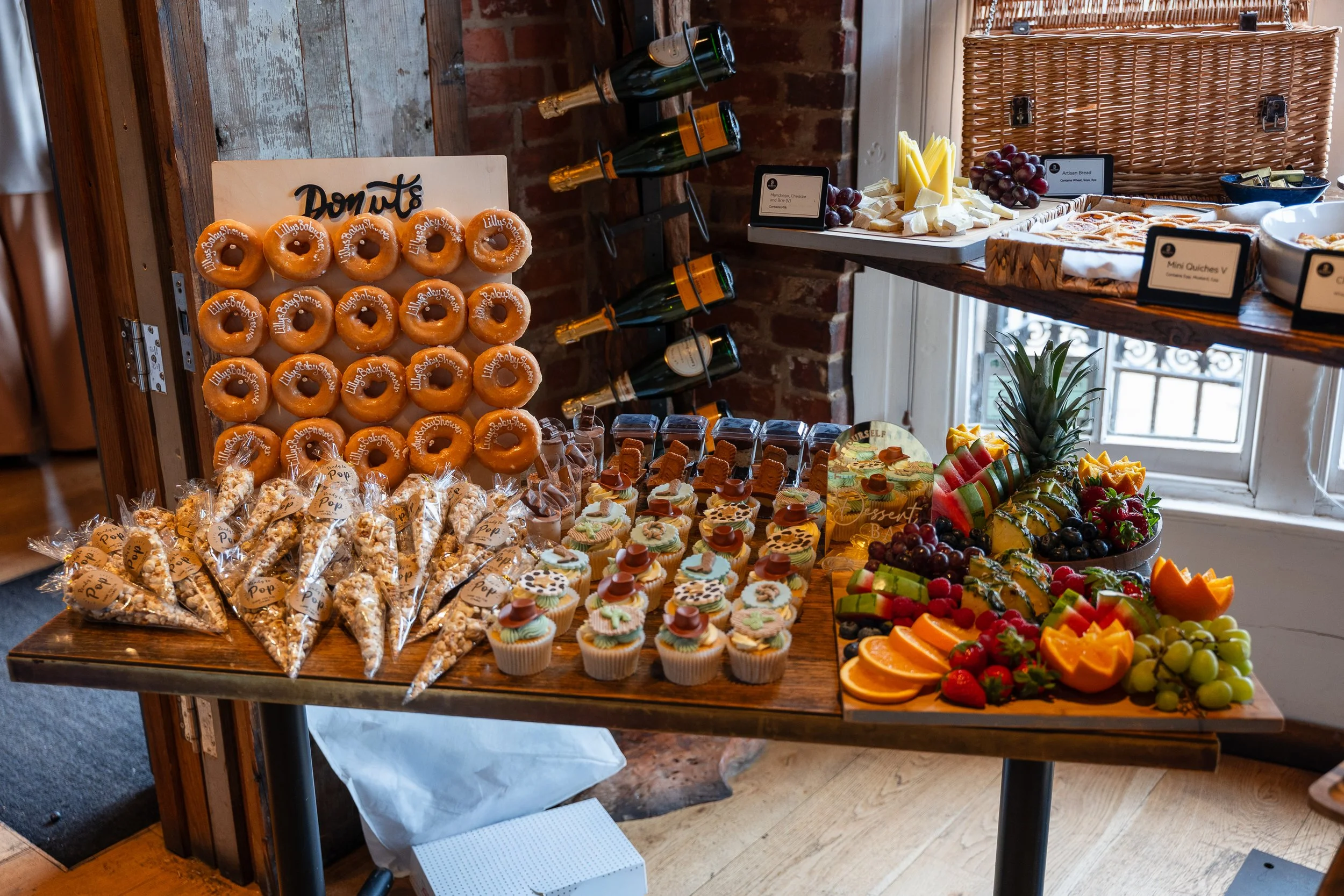 A table set with various baked goods, fruits, and drinks at a buffet or breakfast spread. There are donuts with a sign labeled 'Donuts,' cheese, grapes, watermelon, pineapple, and orange slices, along with other assorted pastries and packaged popcorn snacks. A wicker basket and wine bottles are also visible on the table.
