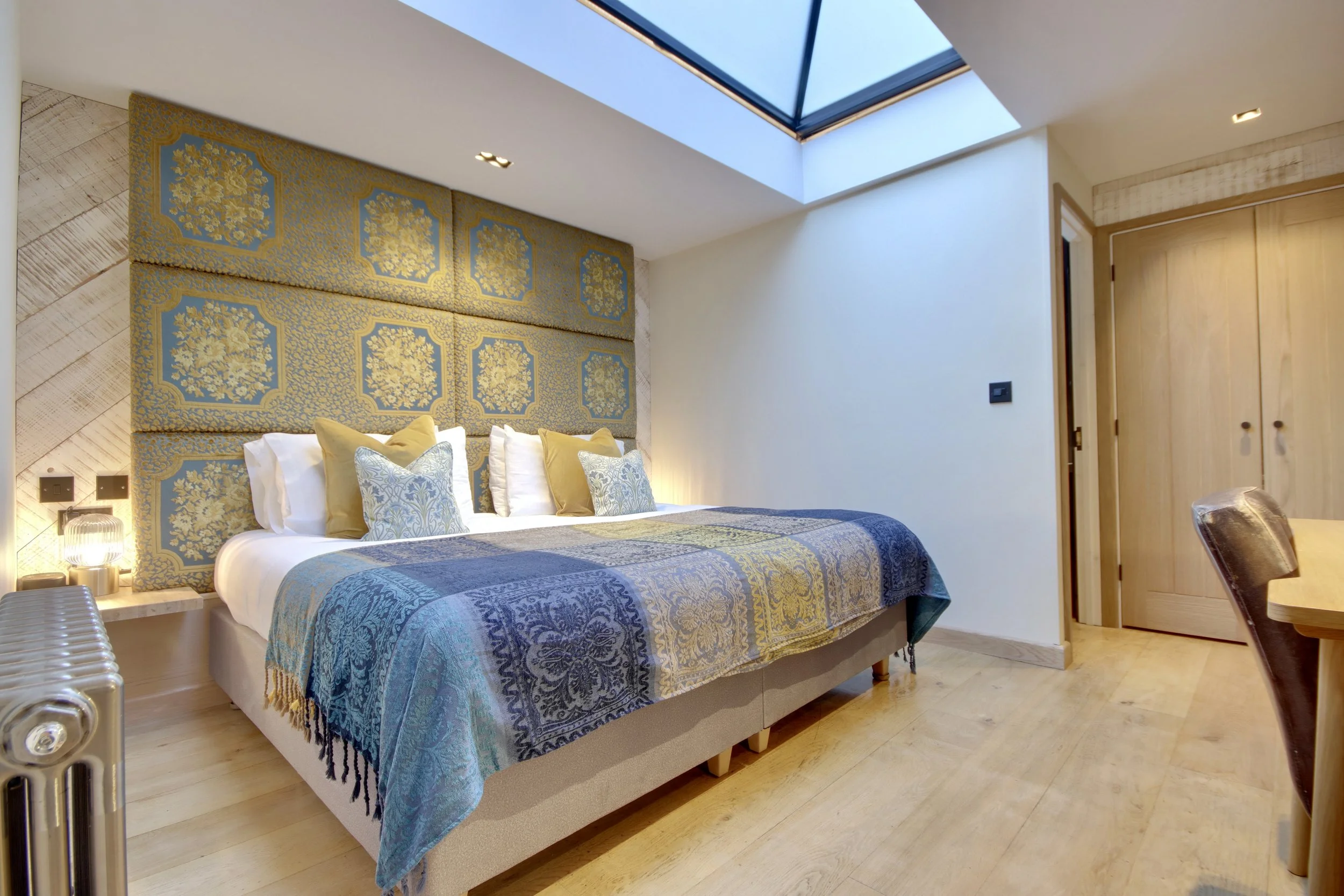 A bedroom with a large bed featuring patterned blue and gold bedding and multiple pillows. The wall behind the bed has a decorative upholstered headboard with a floral pattern. There is a skylight in the ceiling providing natural light. To the right, a wooden closet door and a part of a dining table with a chair are visible.