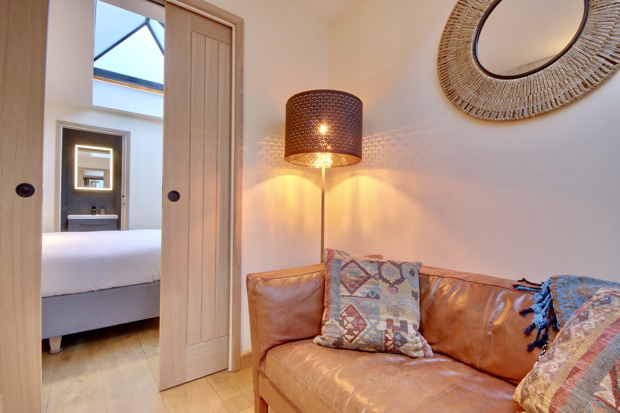 A cozy hotel room with a leather sofa, patterned pillows, a floor lamp, and a round mirror on the wall. A doorway leads to a bedroom with a bed and modern bathroom.