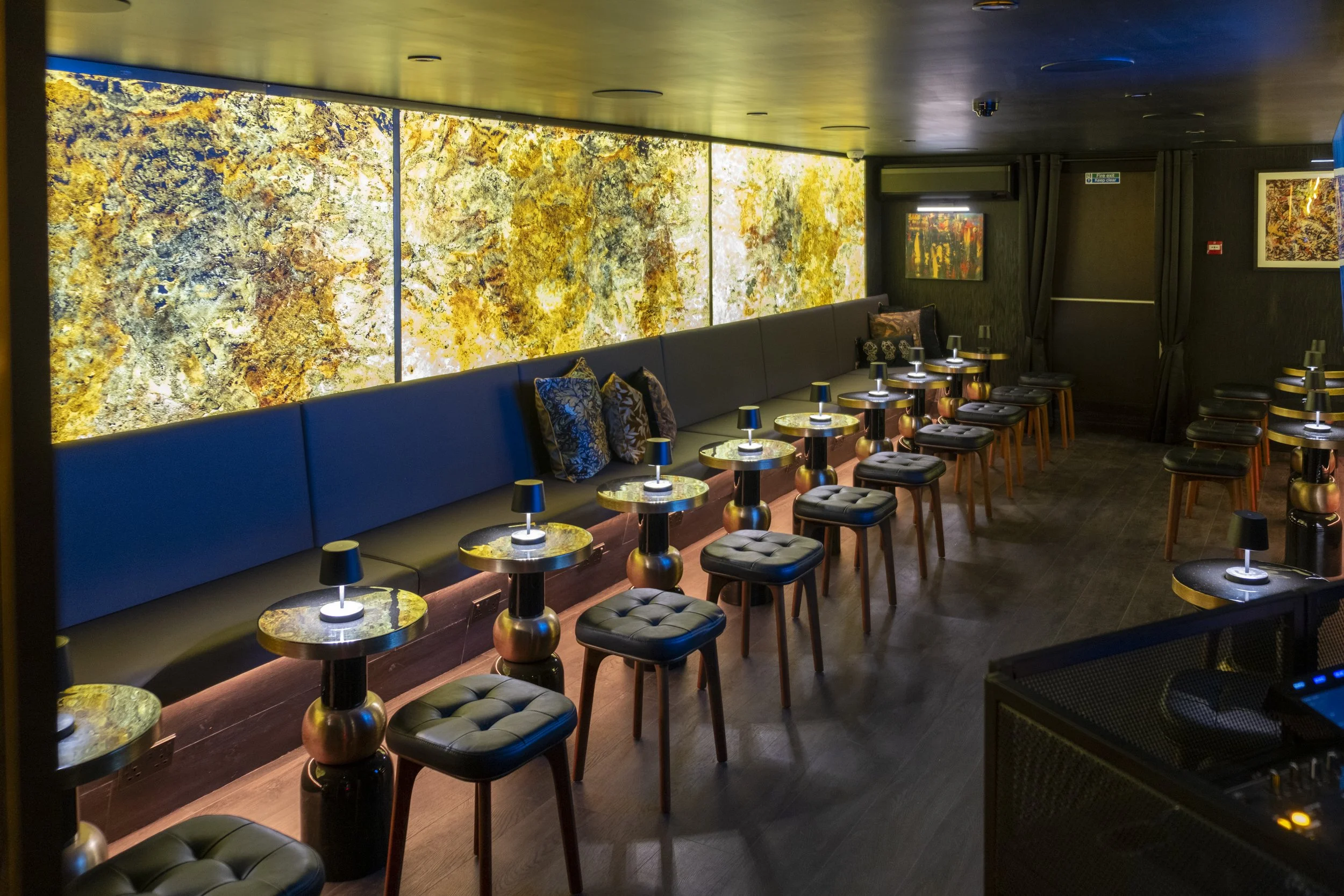 A dimly lit lounge area with a long blue cushioned bench, small round tables with black lamps, and black stools with tufted seats. The background features illuminated textured wall panels with a gold and white pattern, and framed artwork on dark wall
