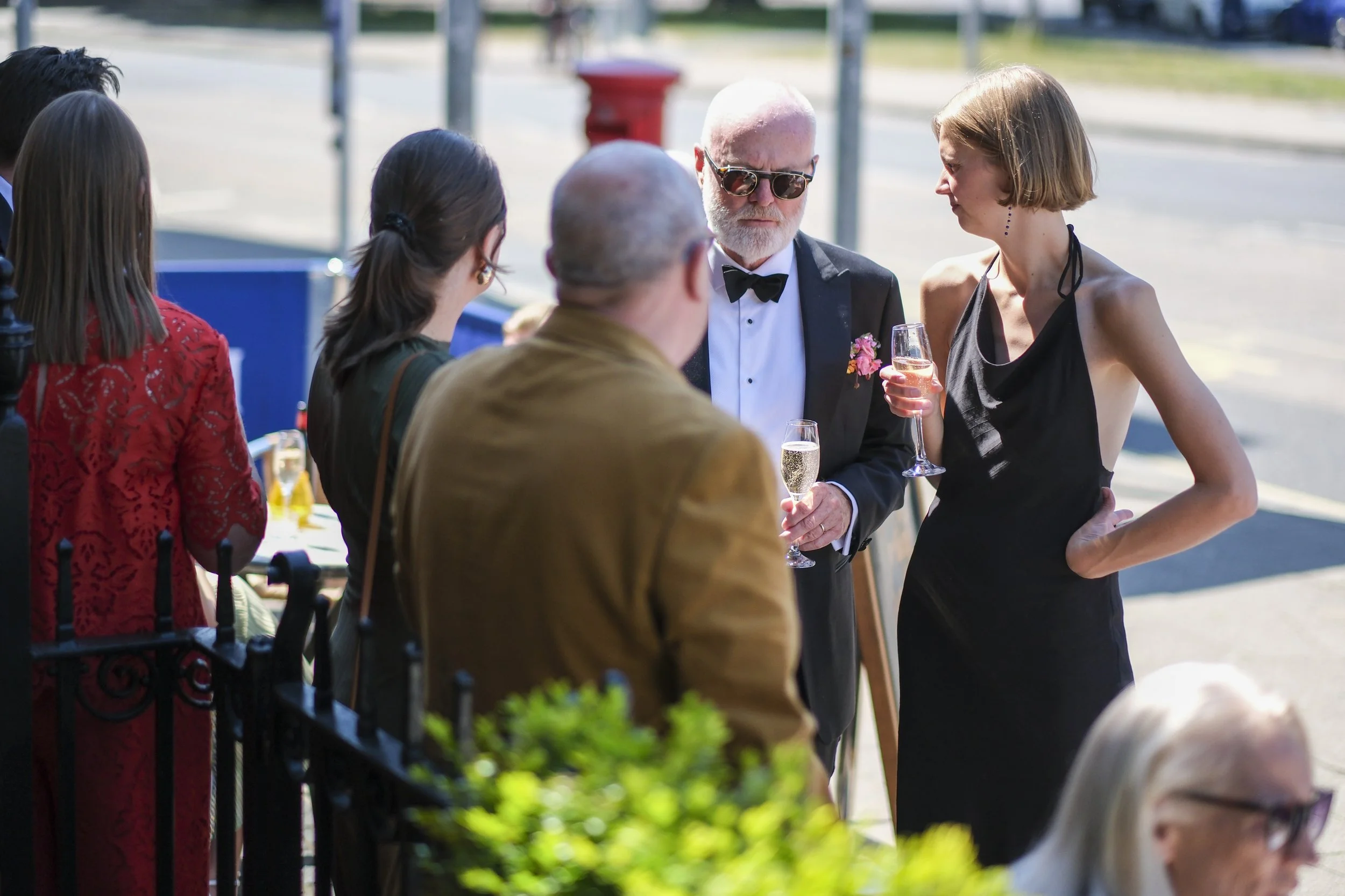 Group of people gathered outdoors, talking, with one man in a tuxedo and sunglasses holding a glass of champagne, and a woman in a black dress holding a glass of champagne, engaging in conversation.