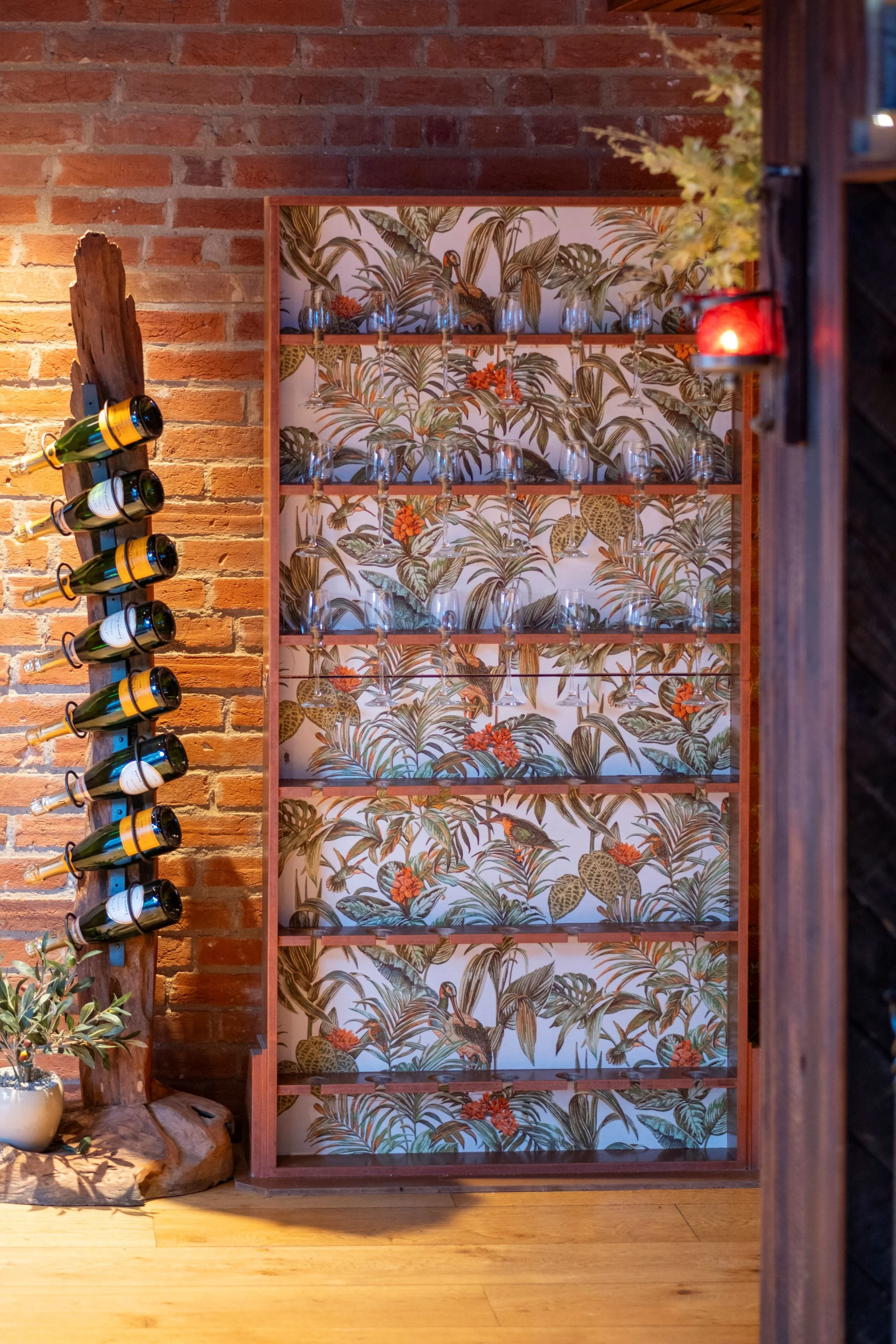 A wooden wine rack with glass holders and a colorful tropical wallpaper background featuring birds, palm leaves, and orange flowers. To the left, mounted wine bottles on a wooden post, and on the lower left, a stone and plant decoration. A small side table is partially visible on the left.