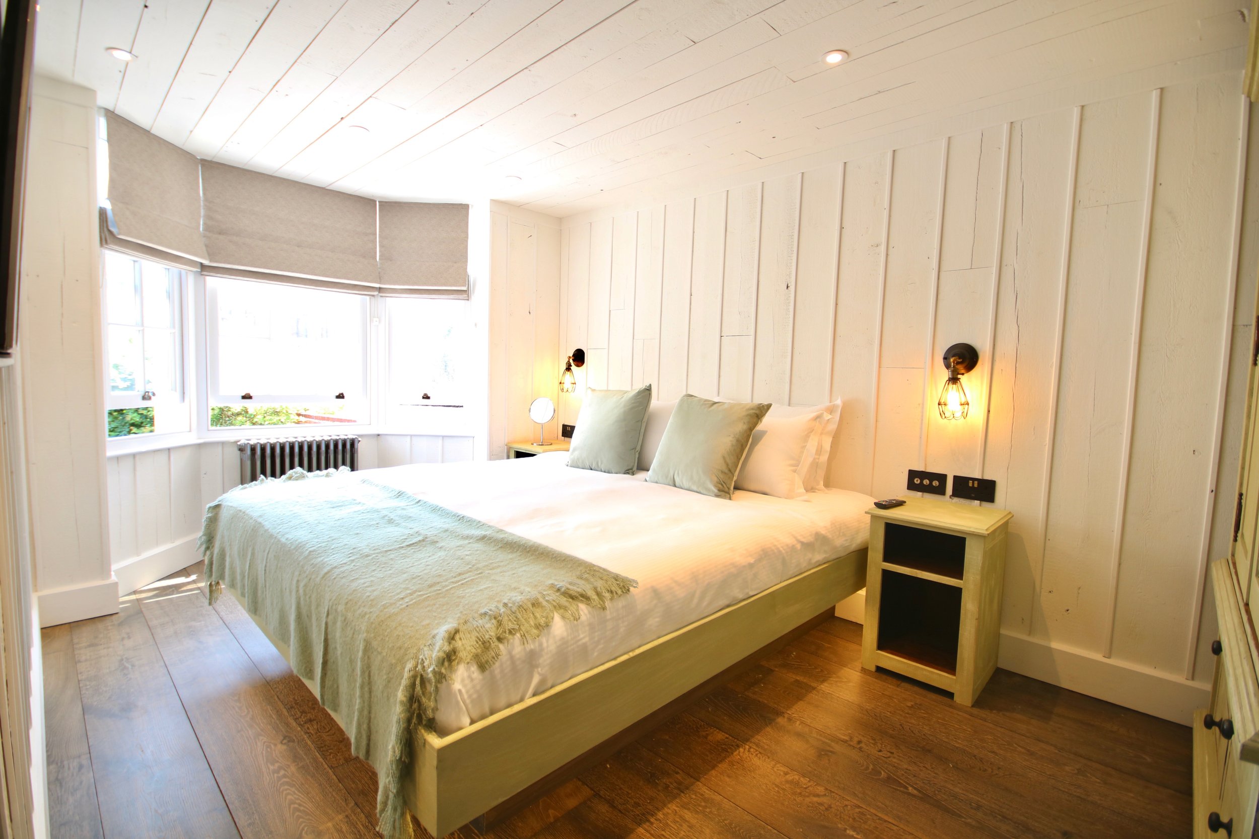 A bedroom with a large bed covered with a beige throw blanket and light-colored pillows, situated next to a window with beige blinds. The room has white wooden panel walls and ceiling, with two black wall-mounted lamps on either side of the bed. There is a small nightstand on the right side with a round lamp and electrical outlets on the wall above. The flooring is dark wood.