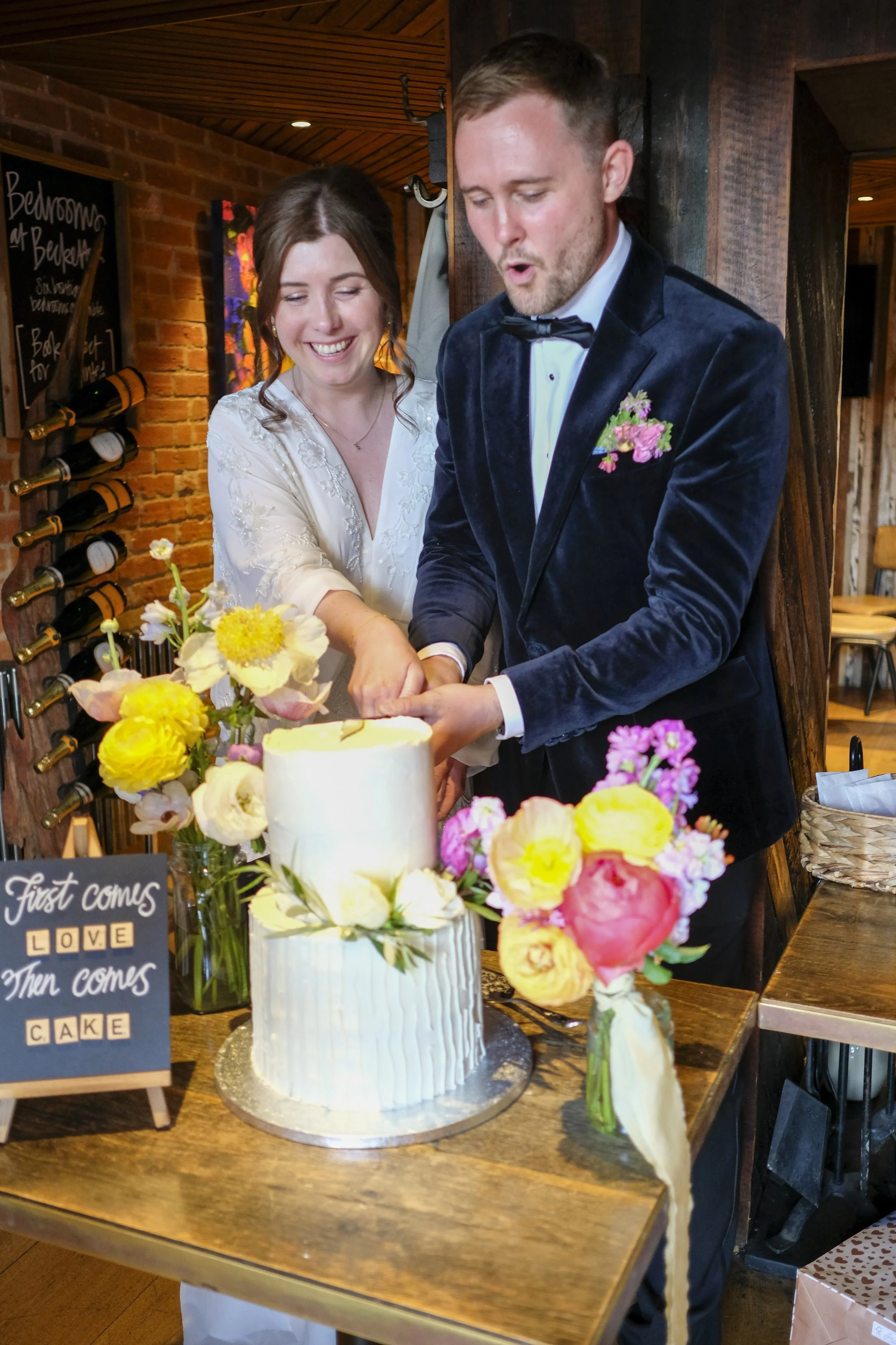 A couple in formal attire cutting a wedding cake at a celebration with flowers on the table.