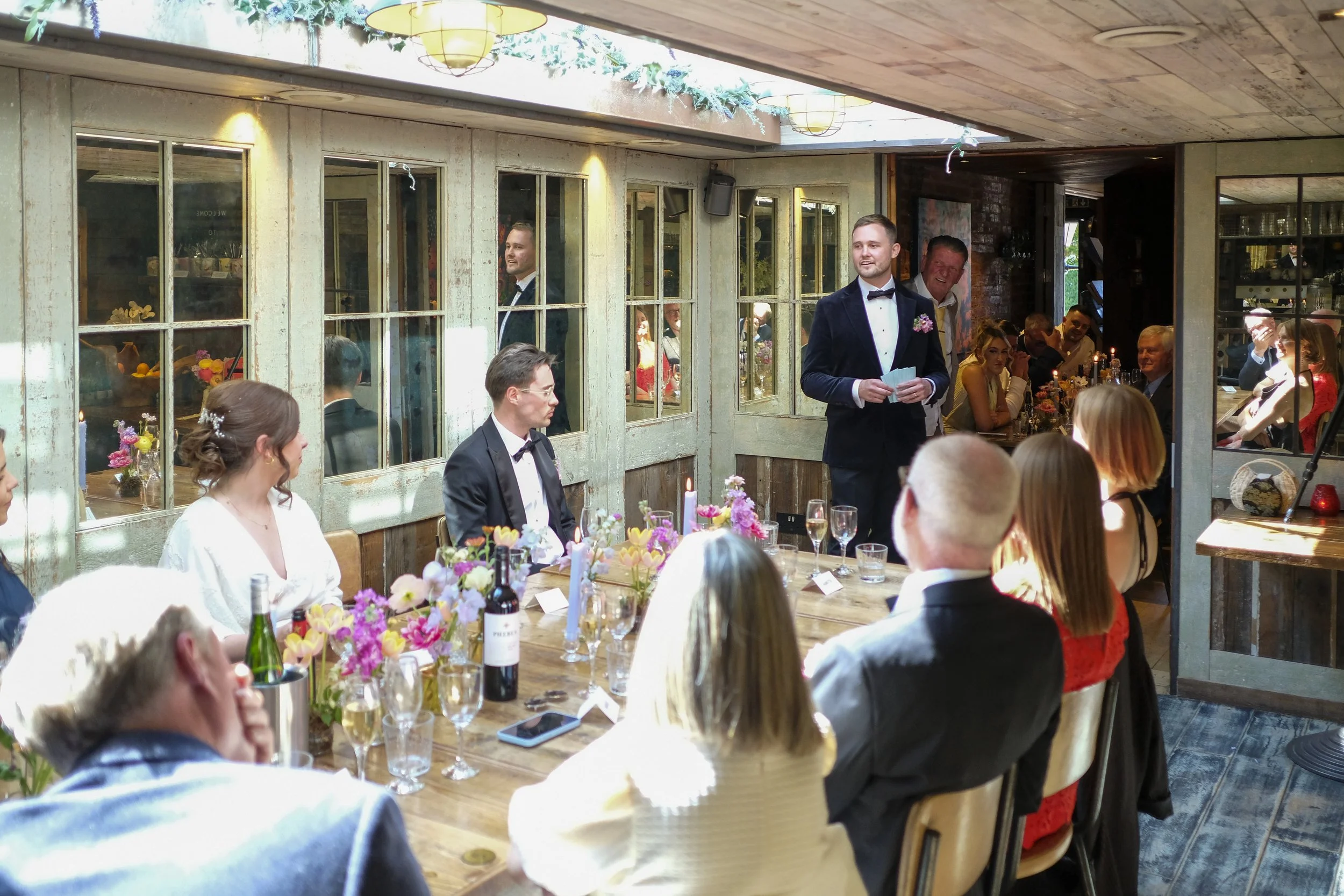A wedding reception with guests seated at tables decorated with flowers, wine bottles, and glasses. A man in a tuxedo is giving a speech while others listen, some smiling and some taking photos in a rustic indoor venue with large windows and wooden d