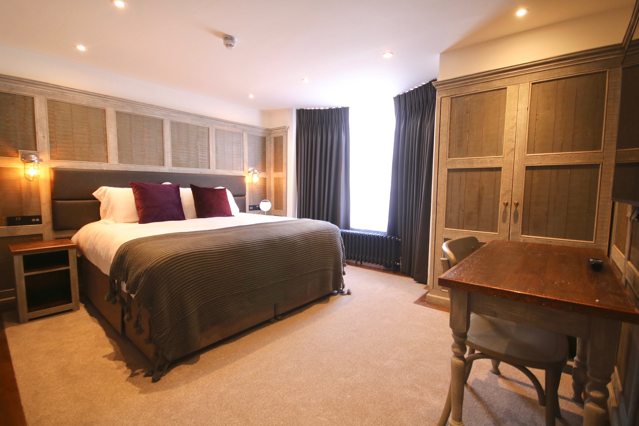 A hotel room with a bed, wooden headboard, purple pillows, nightstand, wall-mounted lamps, a window with dark curtains, a wooden wardrobe, and a wooden table with chairs.