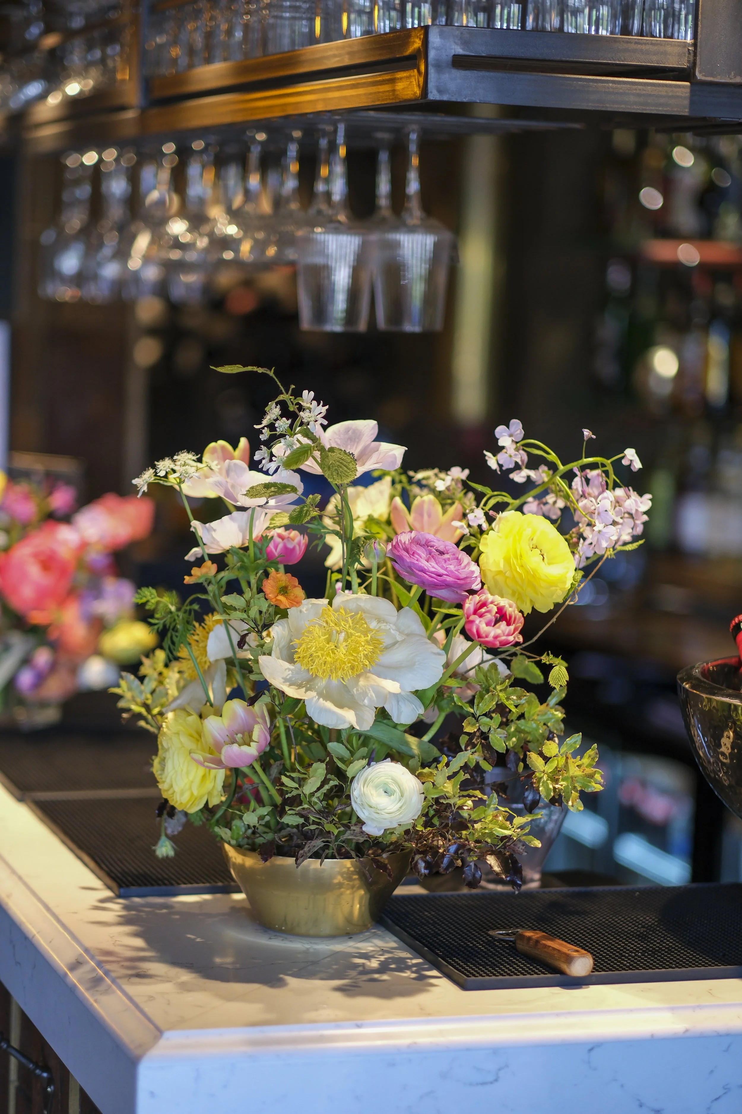 A colorful floral arrangement in a gold vase on a white marble countertop, with blurred shelves and glassware in the background.