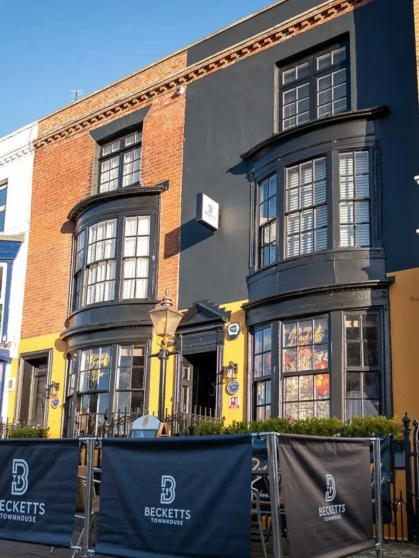 A multi-story building with a colorful facade featuring red brick, black, and yellow sections. It has large bay windows with black trim, decorative wrought iron fencing, and a small outdoor seating area.