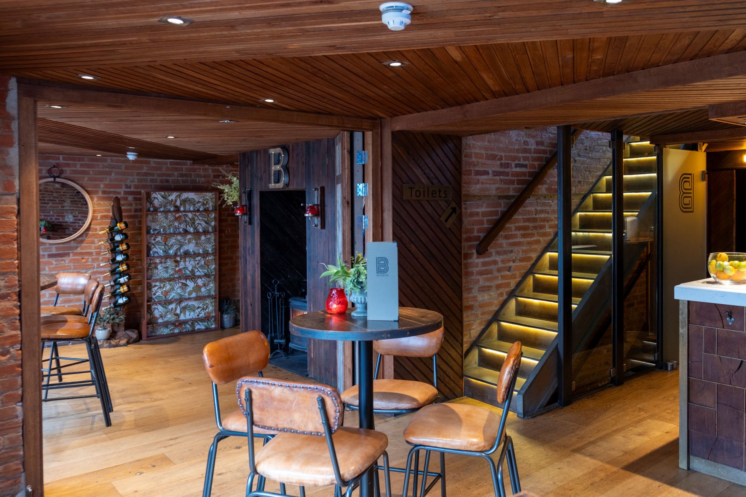 Interior of a restaurant or cafe with wooden ceilings and brick walls, featuring a staircase with circular lighting, a sign pointing to toilets, and seating with brown leather chairs around wooden tables.