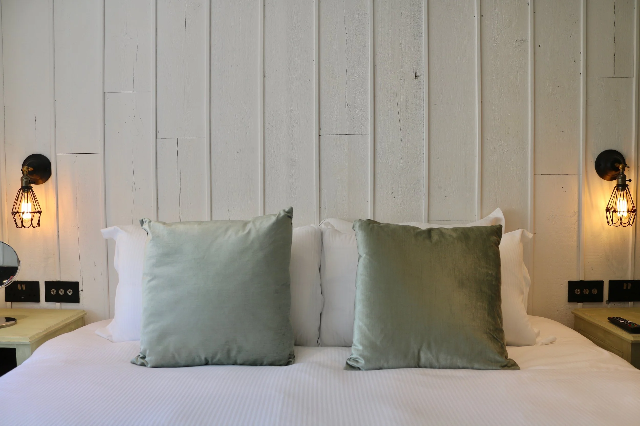 A neatly made bed with white linens and four plush green pillows, with two black wall-mounted lights on either side, in a bedroom with a white wooden paneled wall.