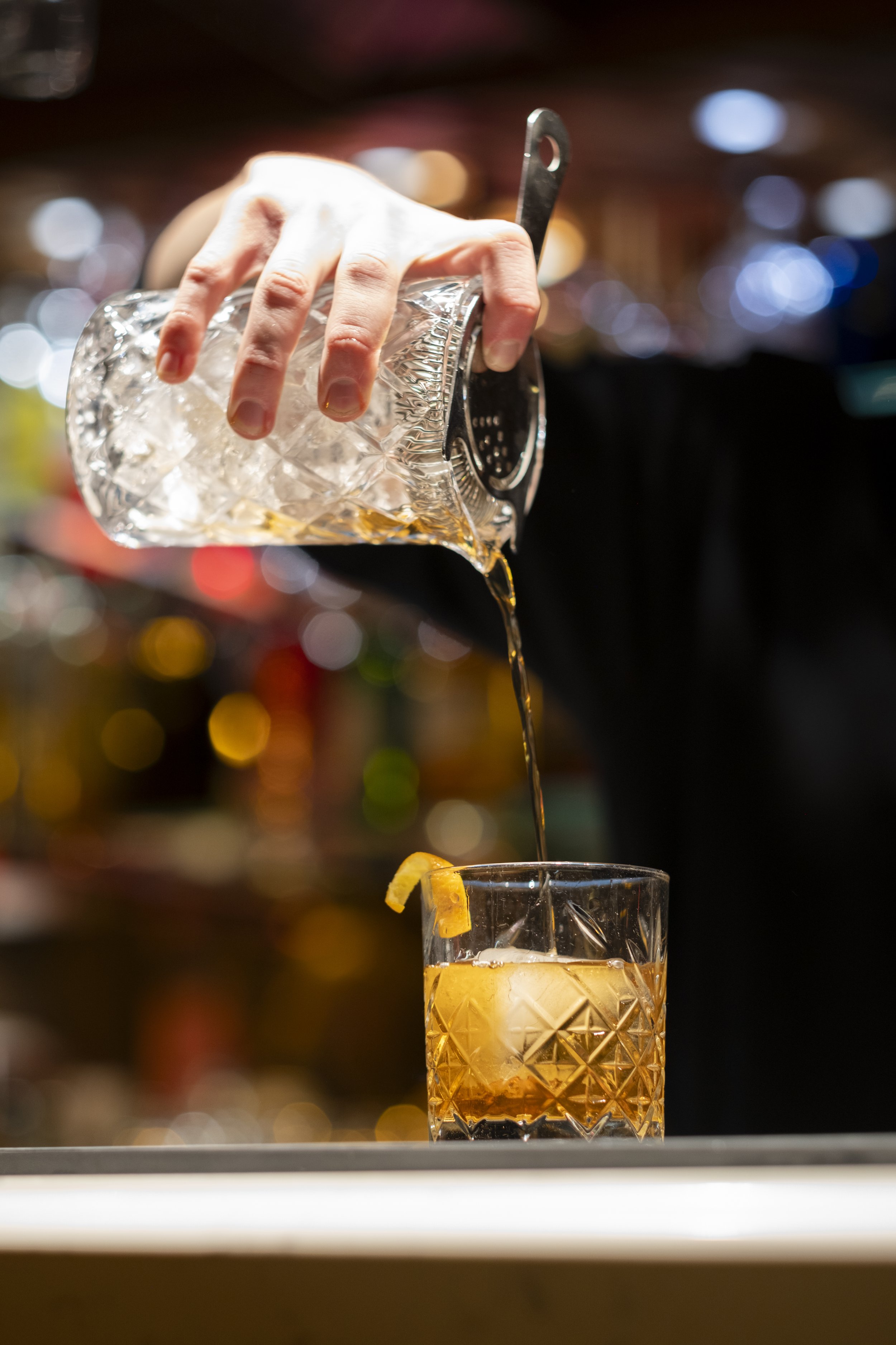 A hand holding a glass mixing jug with ice, pouring a drink into a tall cocktail glass with ice and lemon garnish, with colorful bokeh lights in the background.