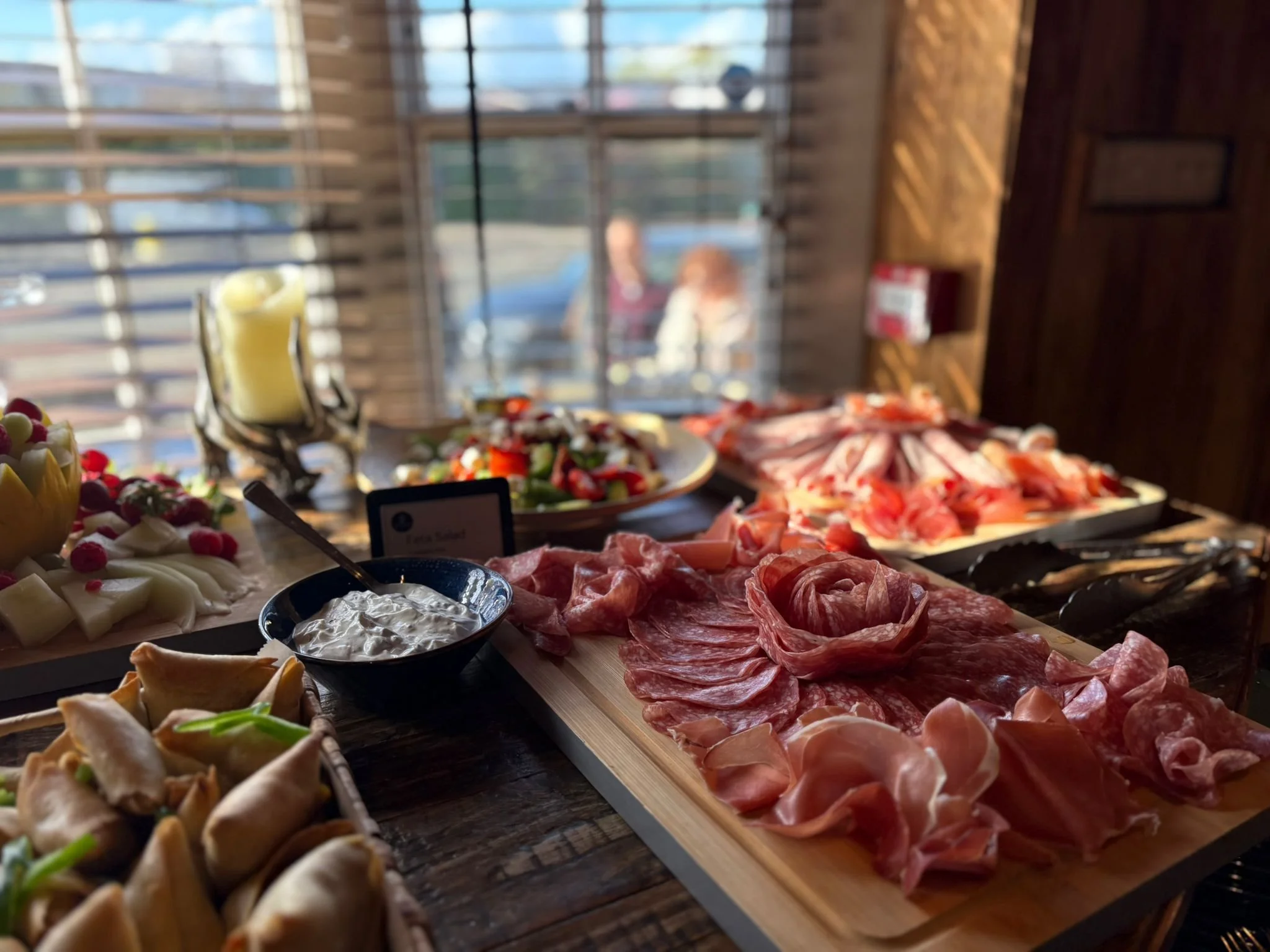 A buffet table with various appetizers including cheeses, meats, salads, and small desserts, with candles and plants in the background.