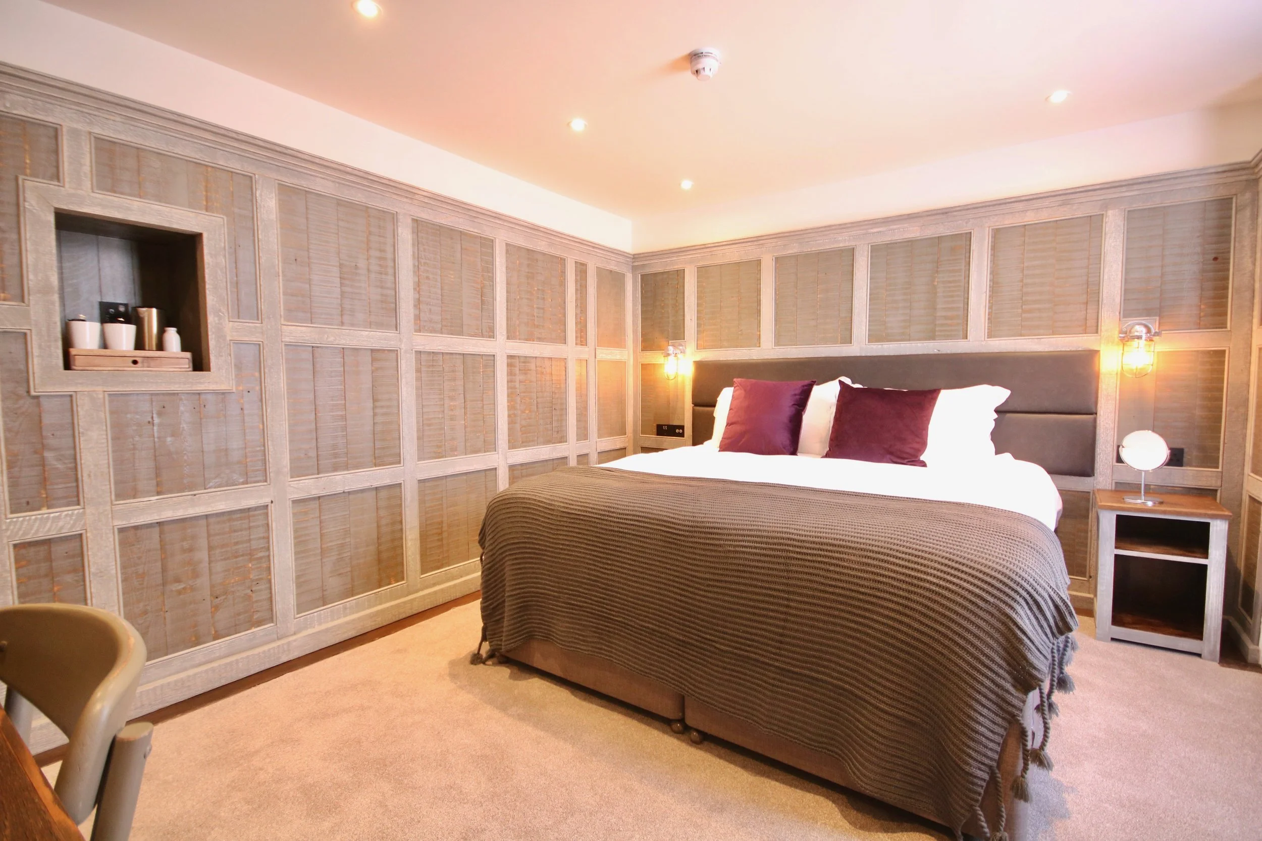 A bedroom with a bed, purple pillows, and a dark headboard. The wall behind the bed has decorative wooden panels with white framing. there are two wall-mounted lamps, a nightstand, and a small spherical lamp. The ceiling has recessed lighting and a smoke detector.