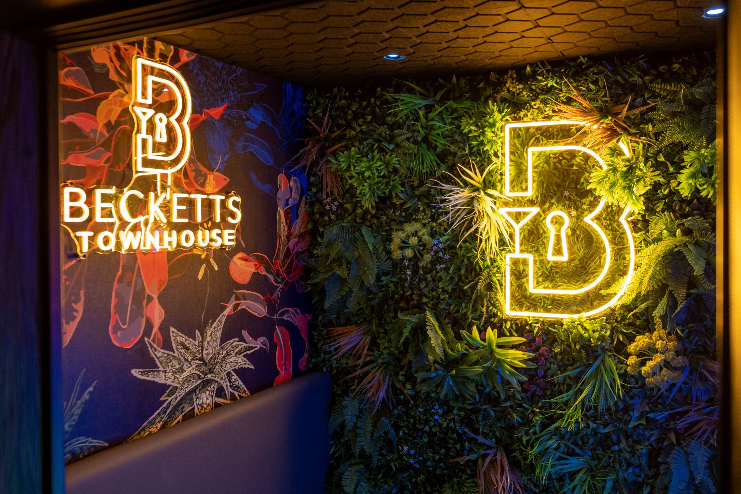 Neon sign and greenery wall at Becketts Townhouse bar, featuring a neon bottle and glass sign, lush green plants, and colorful floral wall art.
