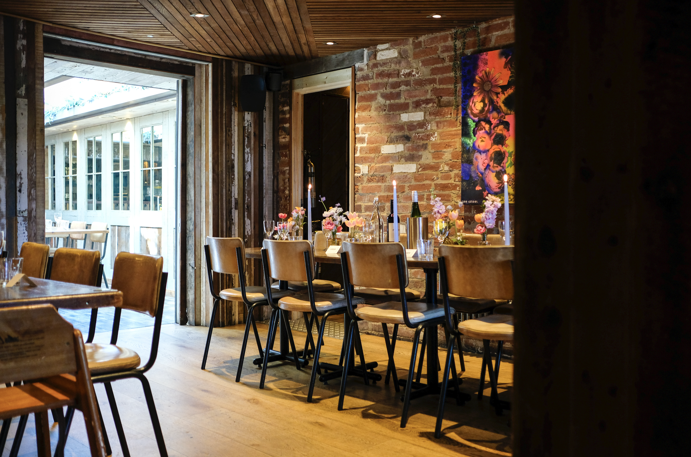 Interior of a cozy dining area with wooden chairs, flower arrangements on the tables, brick walls, and a colorful artwork on the wall. Large windows let in natural light.