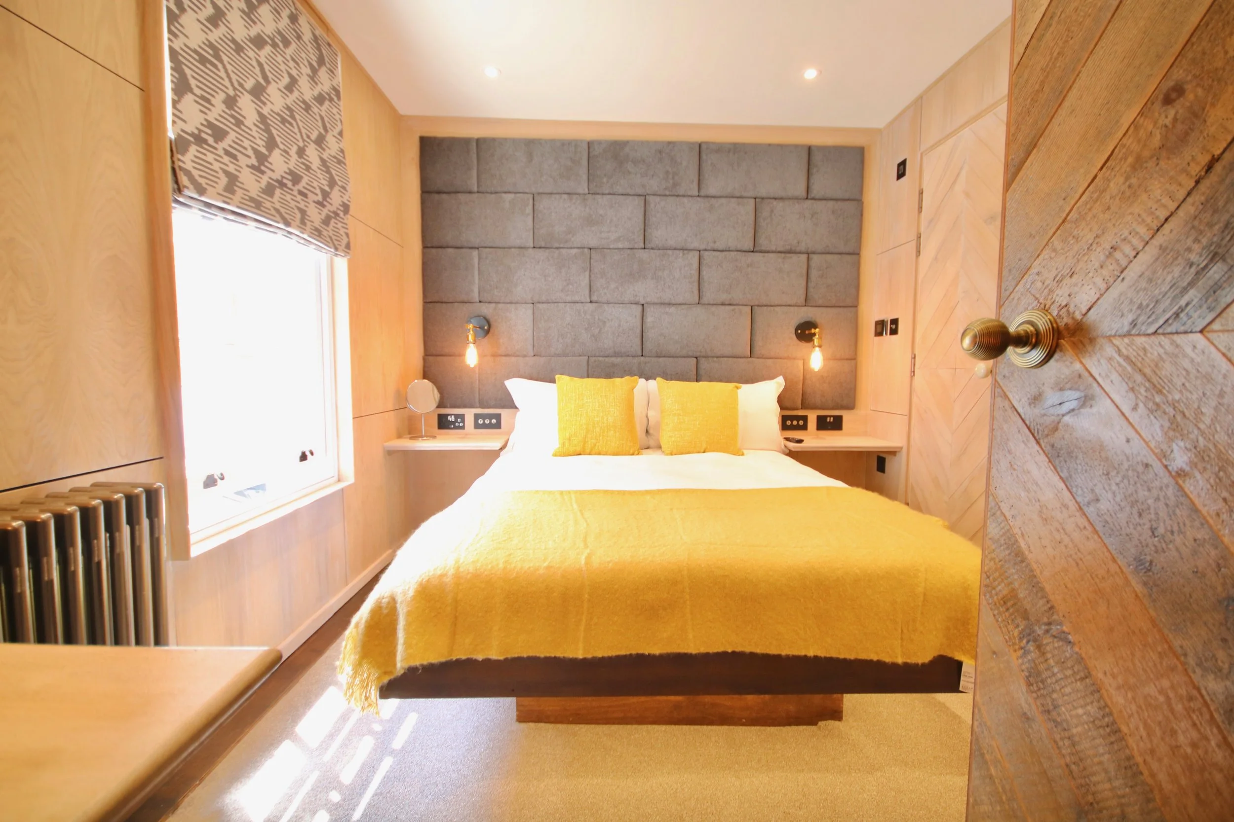 A modern bedroom with a large bed covered in a yellow bedspread and two yellow pillows. The headboard is made of gray upholstered panels. There are two wall-mounted lamps with Edison bulbs on either side of the bed. The room has wooden walls and a window with a patterned blind, allowing sunlight to shine through onto the floor. There is also a radiator and electrical outlets nearby.