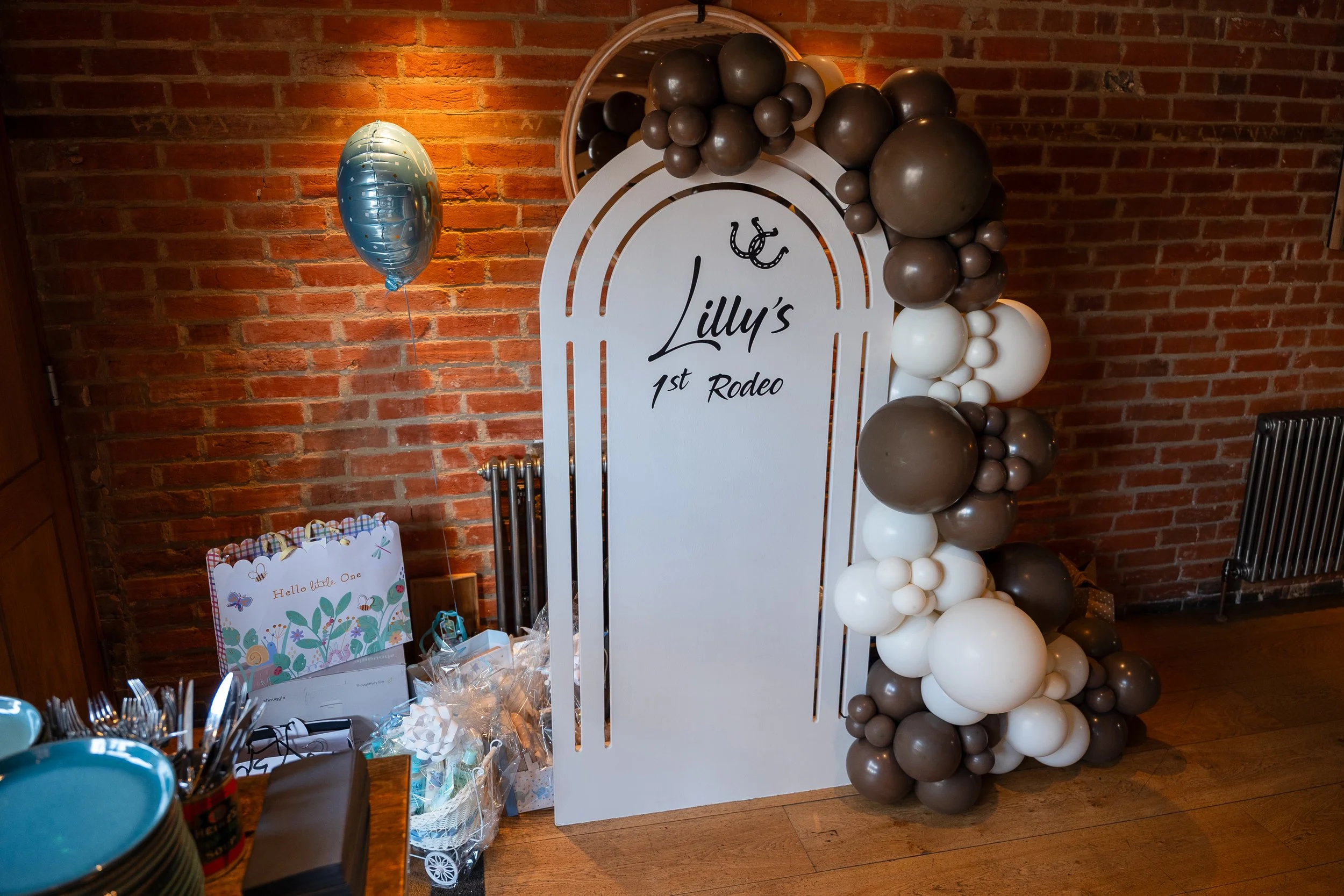 Decorative display for a celebration with a white board reading 'Lilly's 1st Rodeo', decorated with a balloon arch in brown, white, and beige, and a blue balloon with gold writing, set against a brick wall.