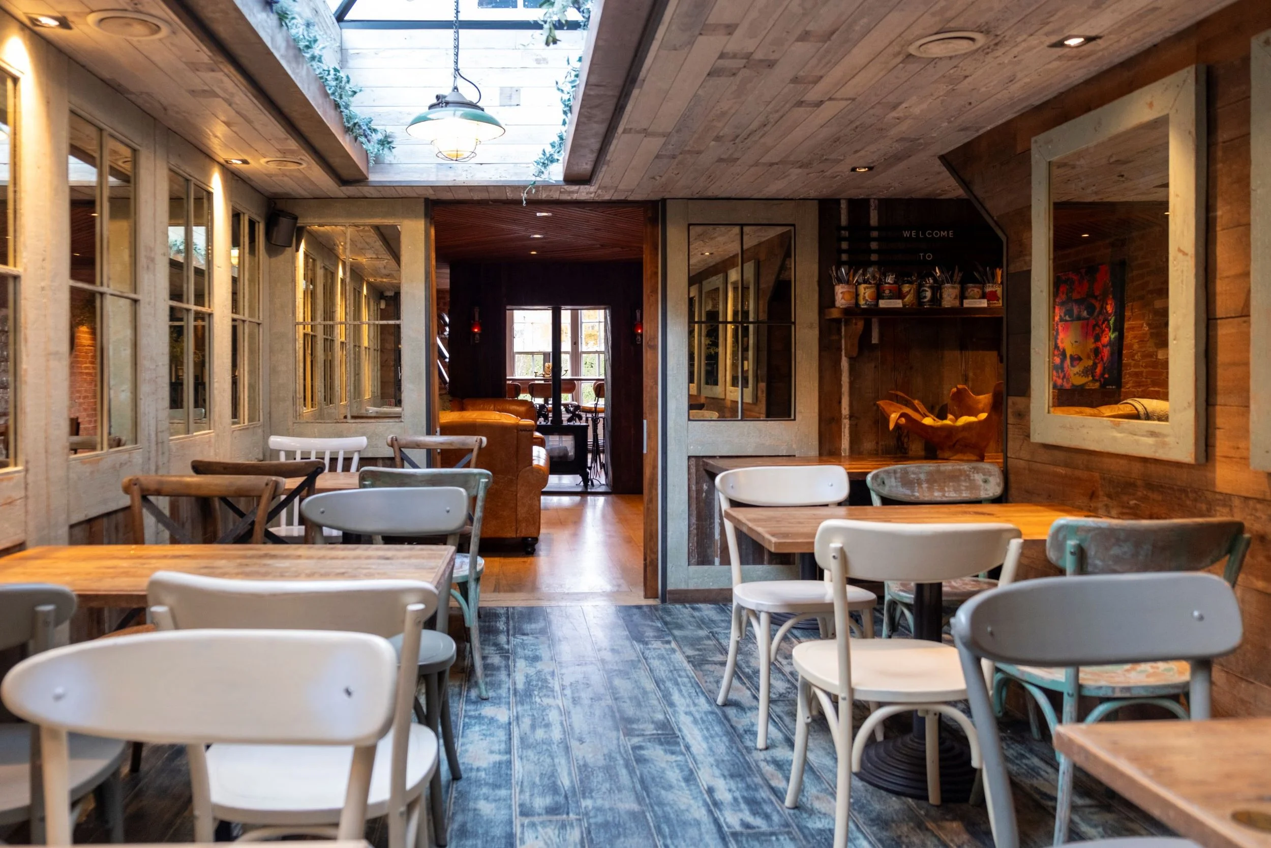 The interior of a cozy restaurant with wooden tables and white chairs, large windows along the wall, a skylight with hanging light fixture, and decorative plants and artwork.