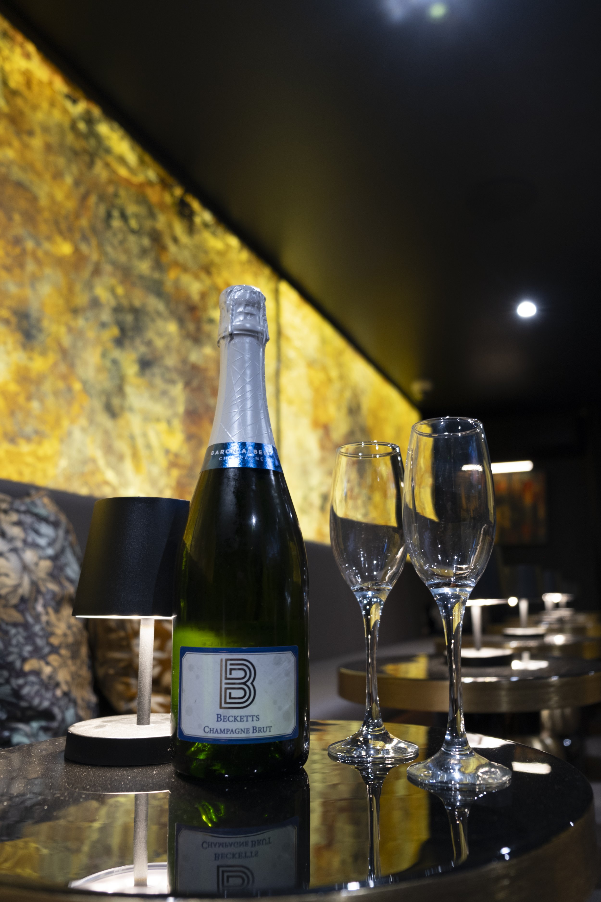 A bottle of Beckett's Champagne Brut with two empty champagne glasses on a reflective black table in a dimly lit restaurant or bar. There is a textured yellow wall in the background and a small table lamp illuminating the scene.