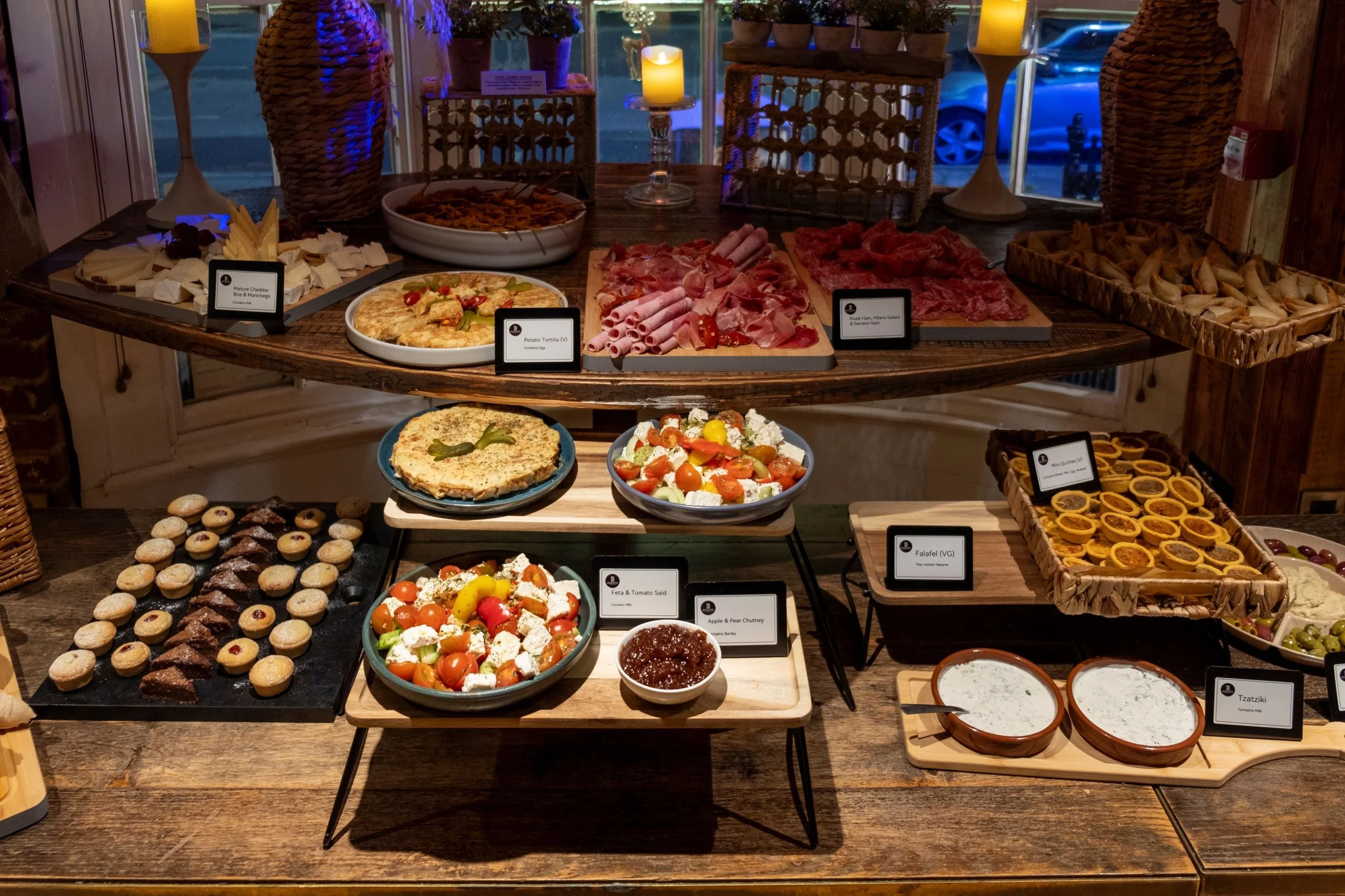 A buffet of various appetizers and snacks displayed on a wooden table, including cheeses, cured meats, salads, mini tarts, and dips, with labels identifying some of the items.