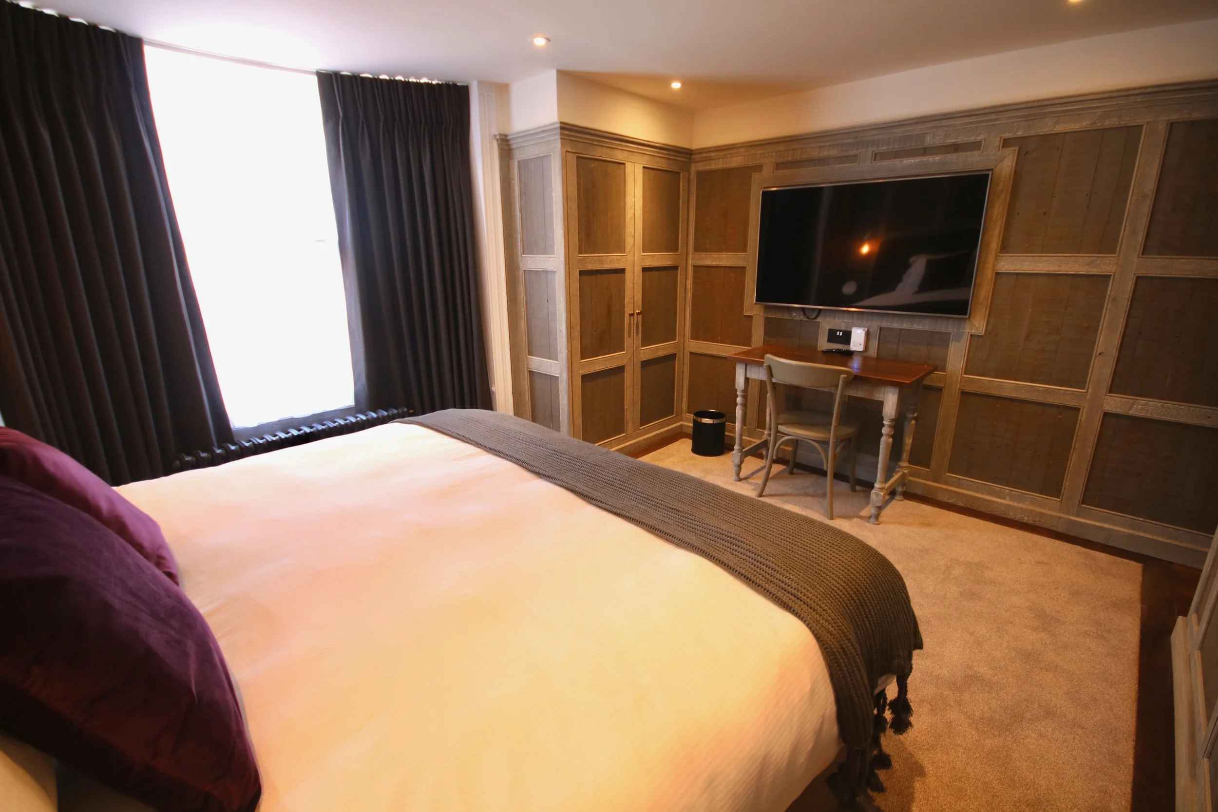 A bedroom with a large bed having a beige bedspread and dark purple pillows, dark curtains on a window, a wooden desk with a chair, a mounted flat-screen TV, and wood-paneled wall decor.